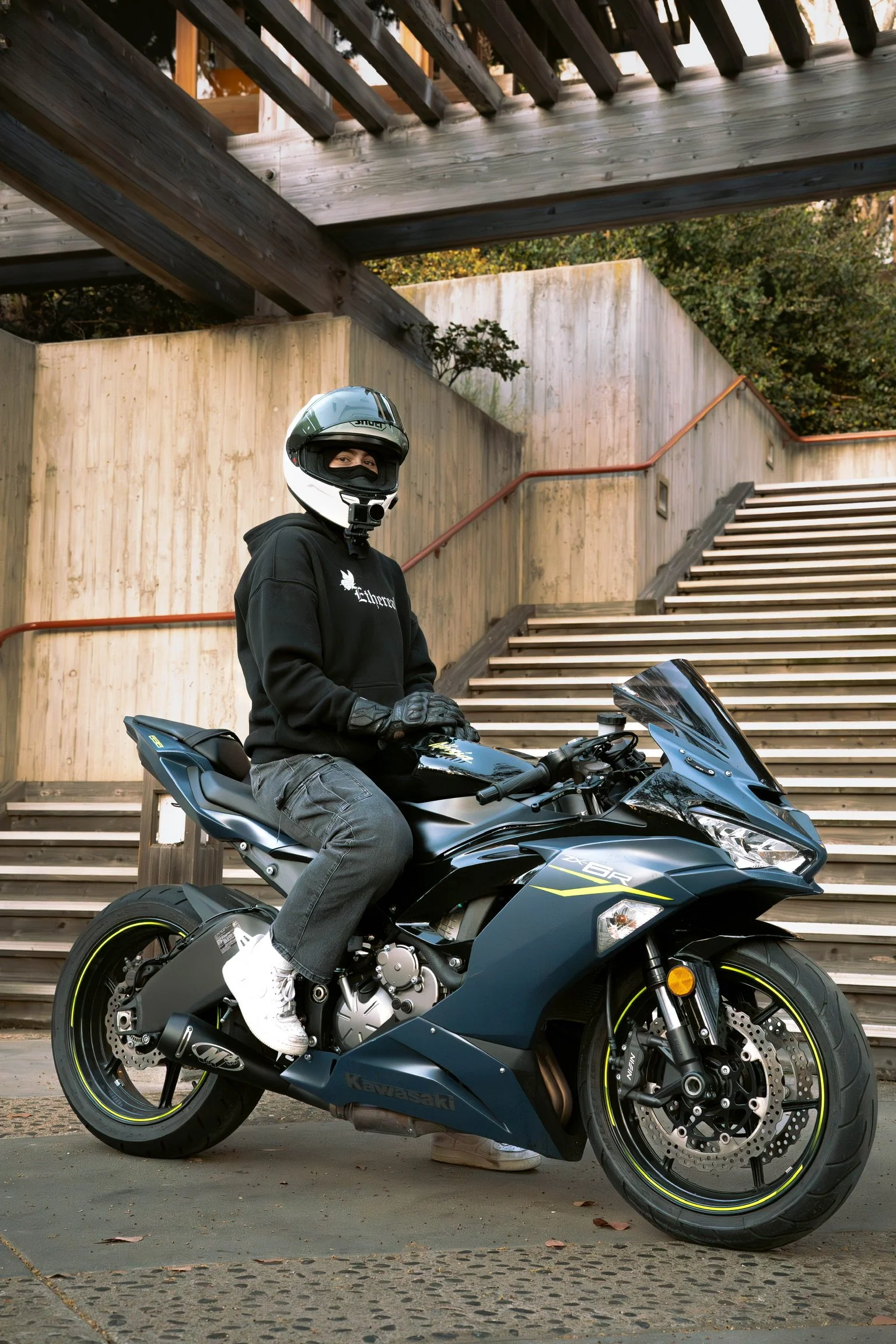 A person wearing a black hoodie, gray jeans, white sneakers, gloves, and a motorcycle helmet is sitting on a black Kawasaki sport motorcycle. The background features wooden stairs and walls, with some greenery above.