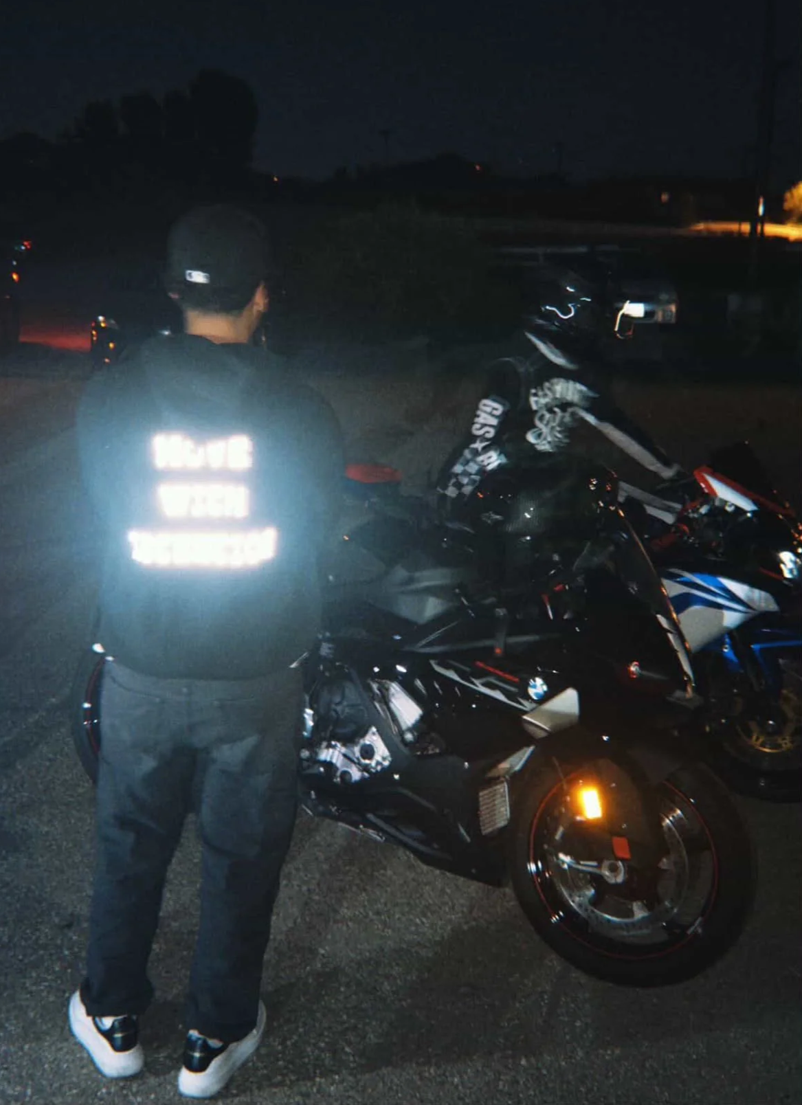 Person wearing a helmet and a reflective vest standing near a sports motorcycle at night in an outdoor parking lot.