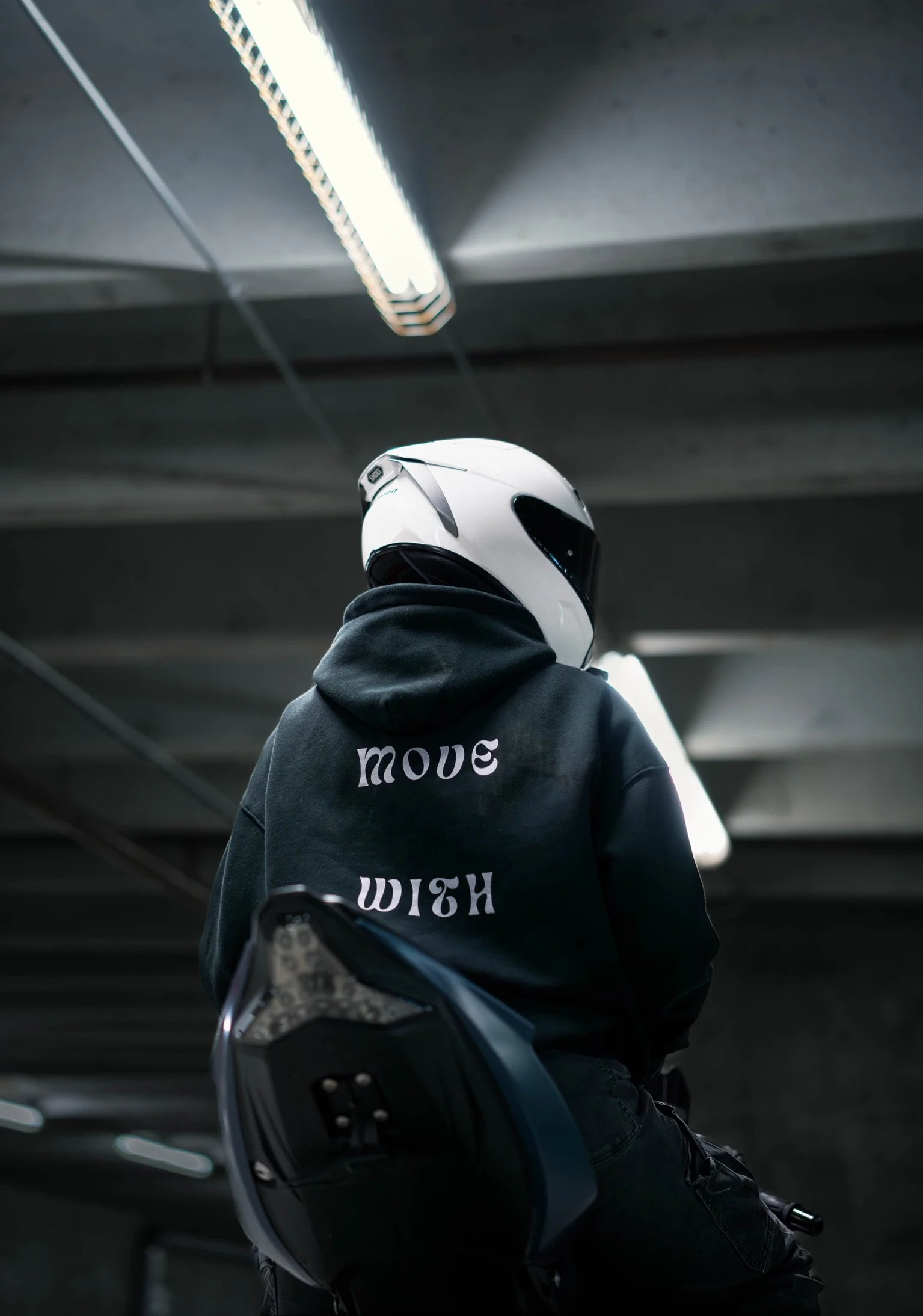 Person wearing a white motorcycle helmet and black hoodie with the words 'move' and 'truth' written on the back, sitting on a dark scooter in an indoor parking garage.