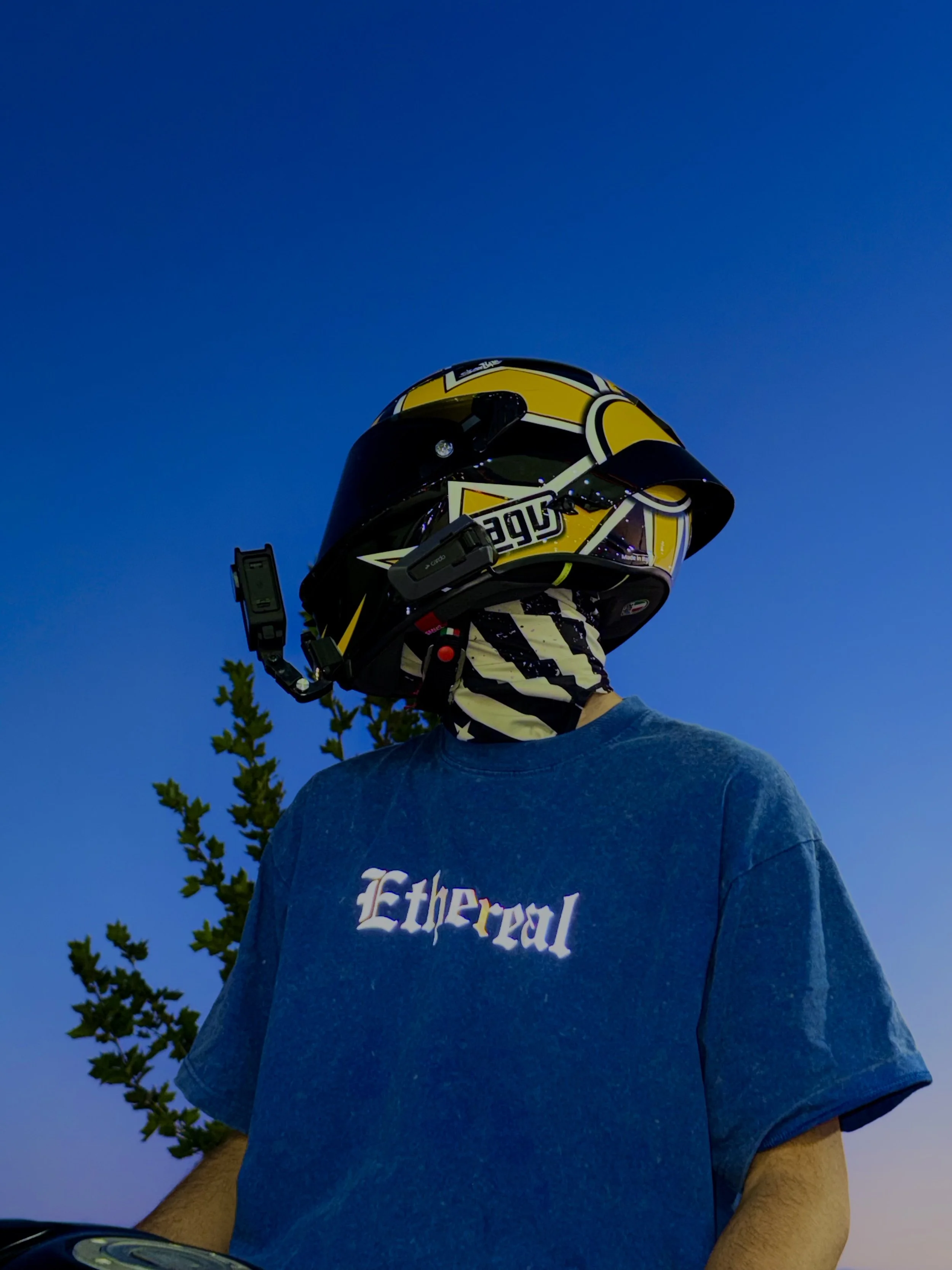 A person wearing a blue T-shirt and a yellow helmet standing outdoors against a clear blue sky with trees in the background.