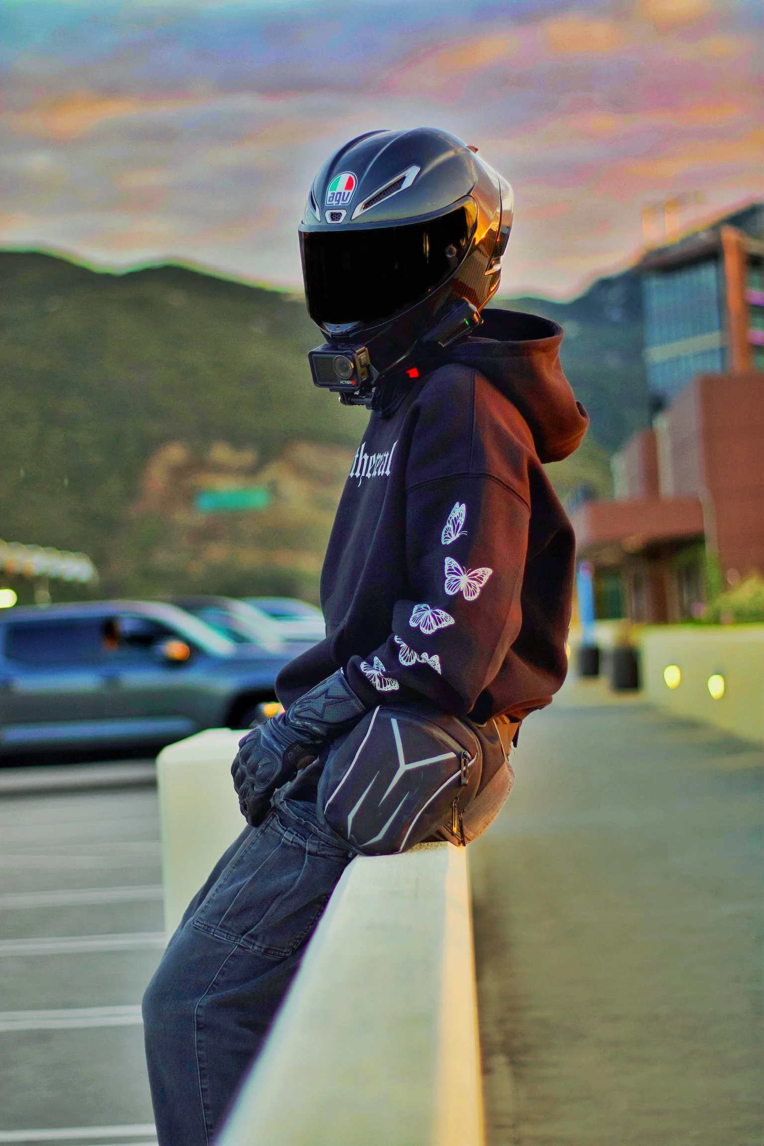 Person leaning against a concrete barrier in a parking lot at sunset, wearing a black helmet with a sticker, a black hoodie with butterfly designs, and gloves, with mountains and buildings in the background.