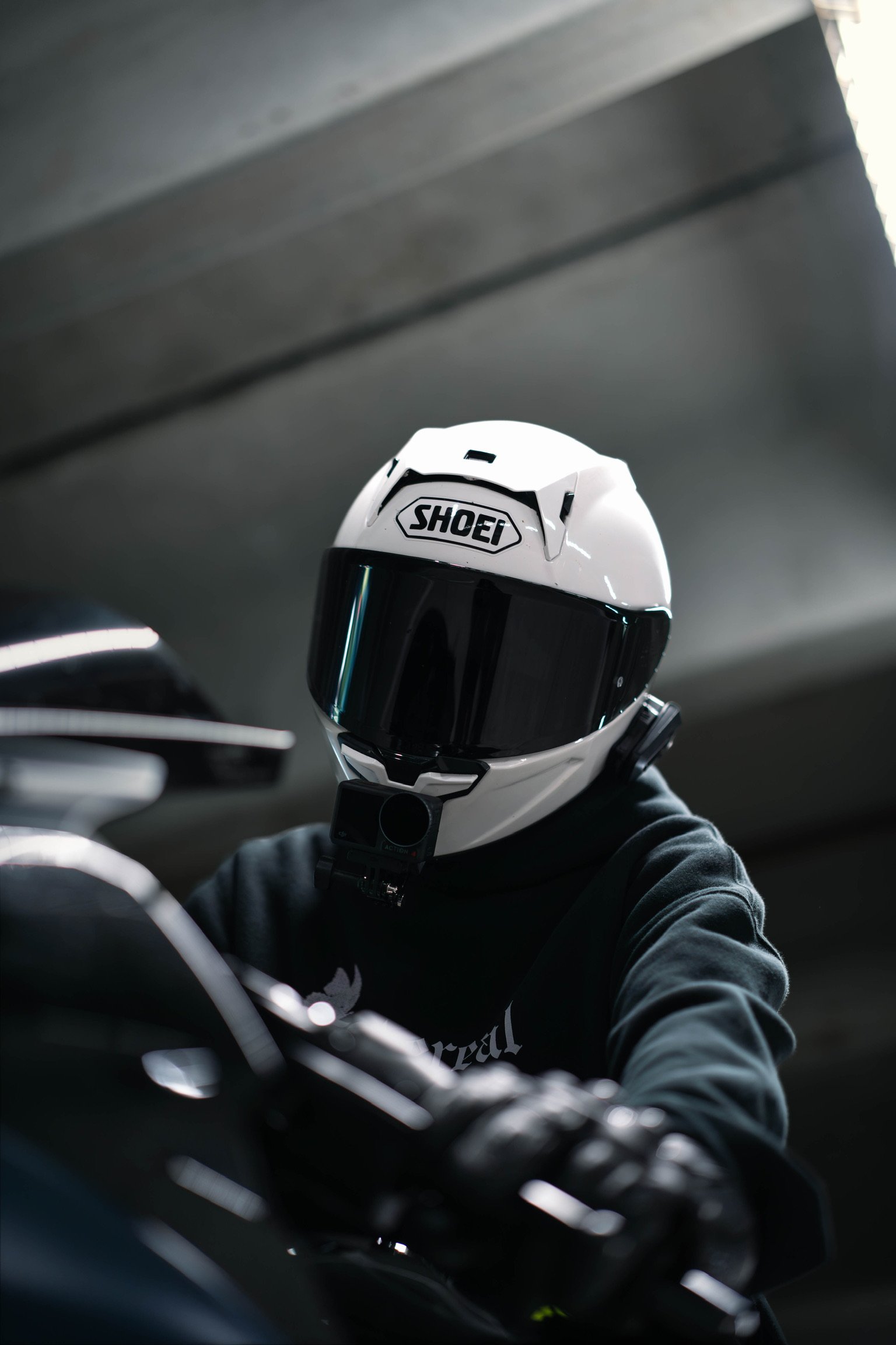 A person wearing a white Shoei motorcycle helmet with a dark visor, seated in a vehicle, with part of a steering wheel and a black hoodie visible. The background appears to be the interior of a vehicle with a blurred, gray ceiling.