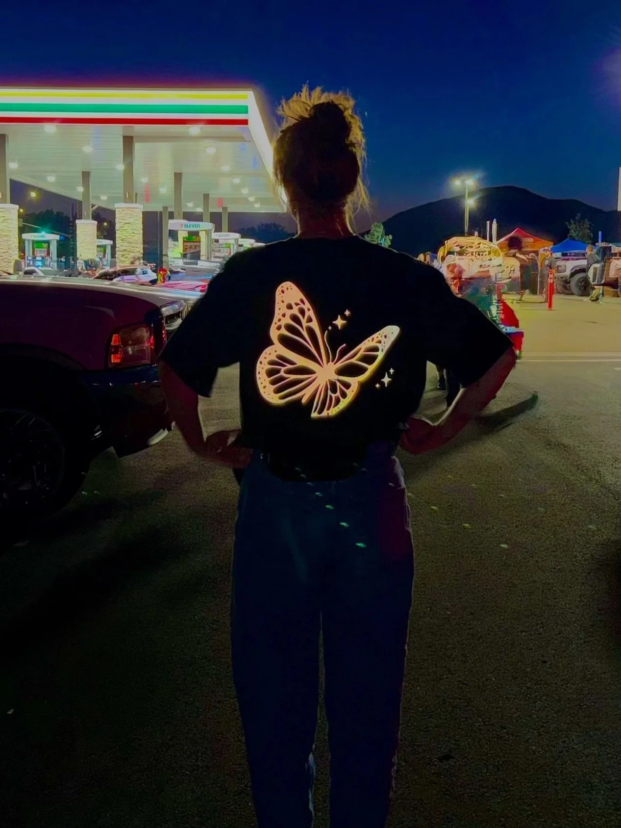 Person with a butterfly design on their shirt standing in a parking lot at night near a 7-Eleven and a gas station.