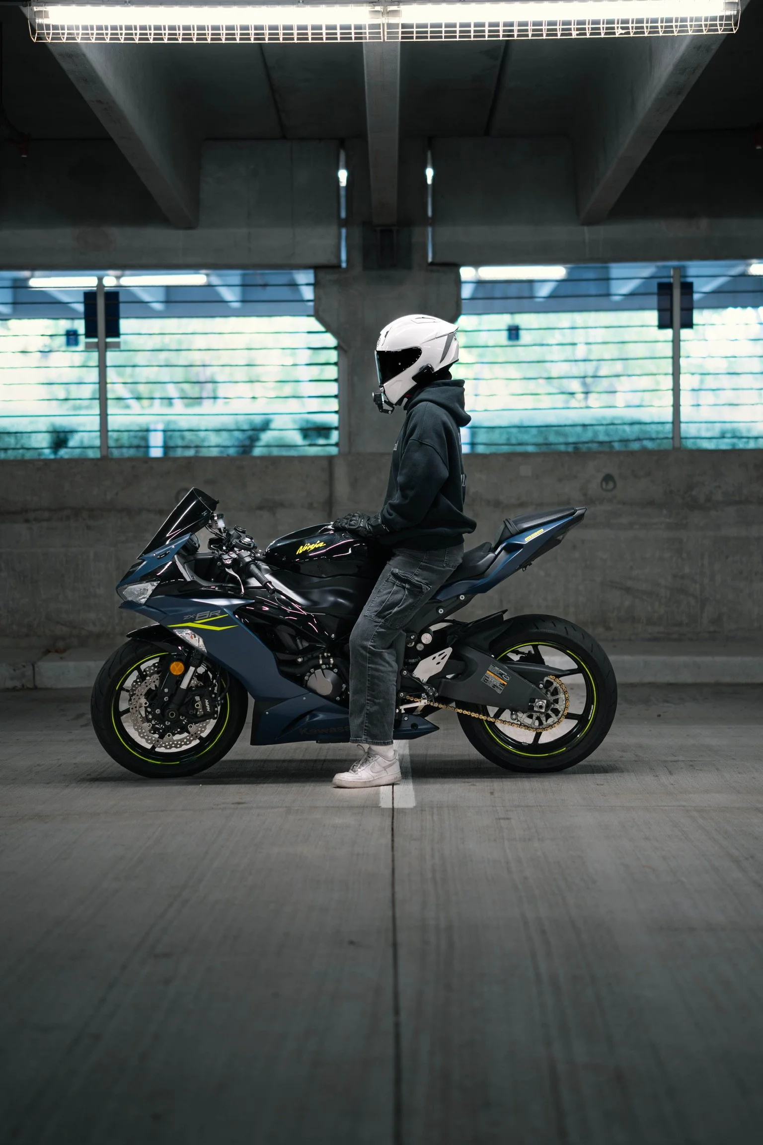 A person wearing a white motorcycle helmet, black hoodie, and gray jeans sitting on a black and blue Kawasaki Ninja motorcycle in an indoor parking garage with concrete walls and ceiling.