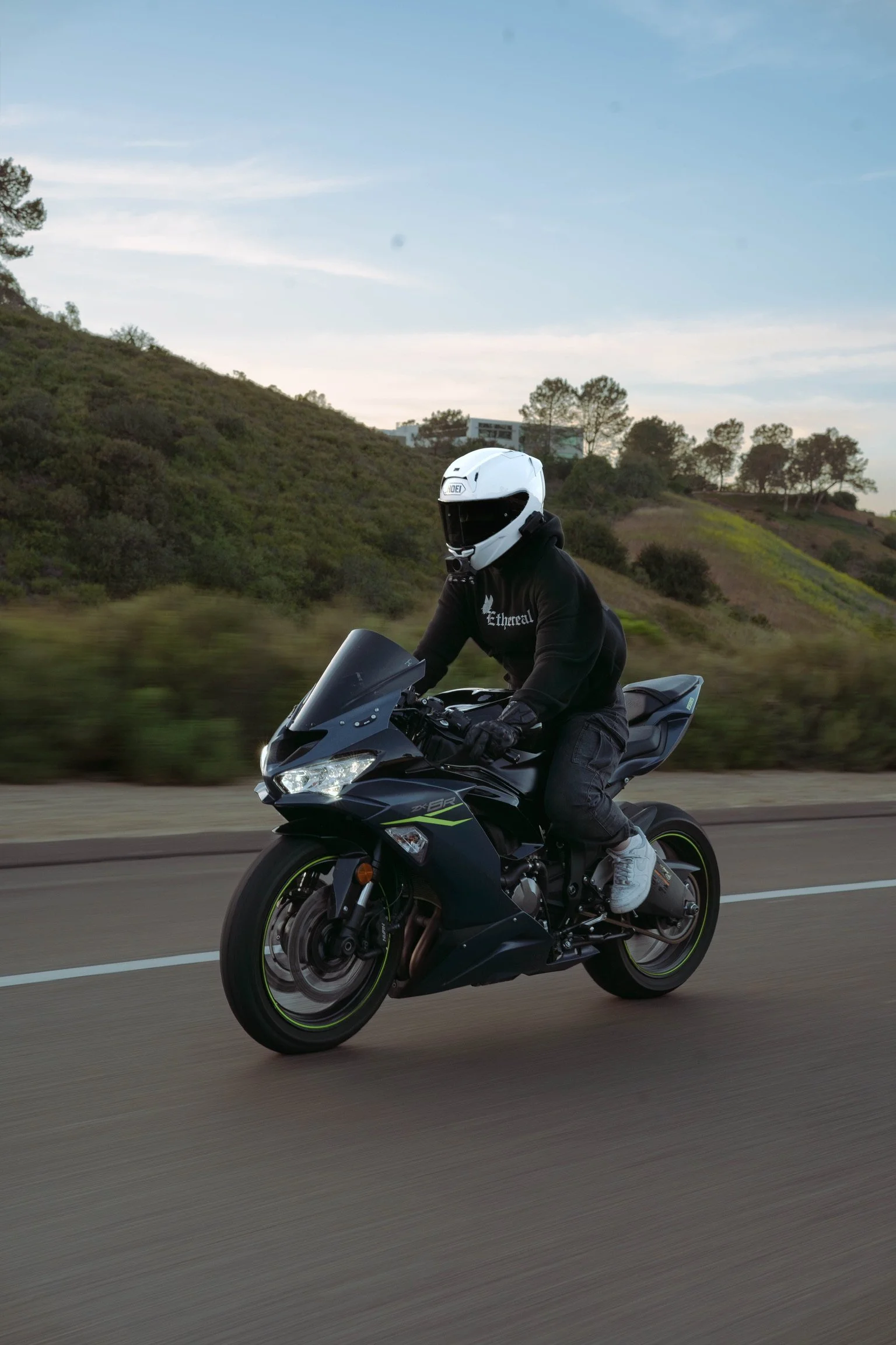 A person riding a black and green Kawasaki sport motorcycle on a road, wearing a white helmet and black hoodie with white lettering, with green hills and trees in the background.
