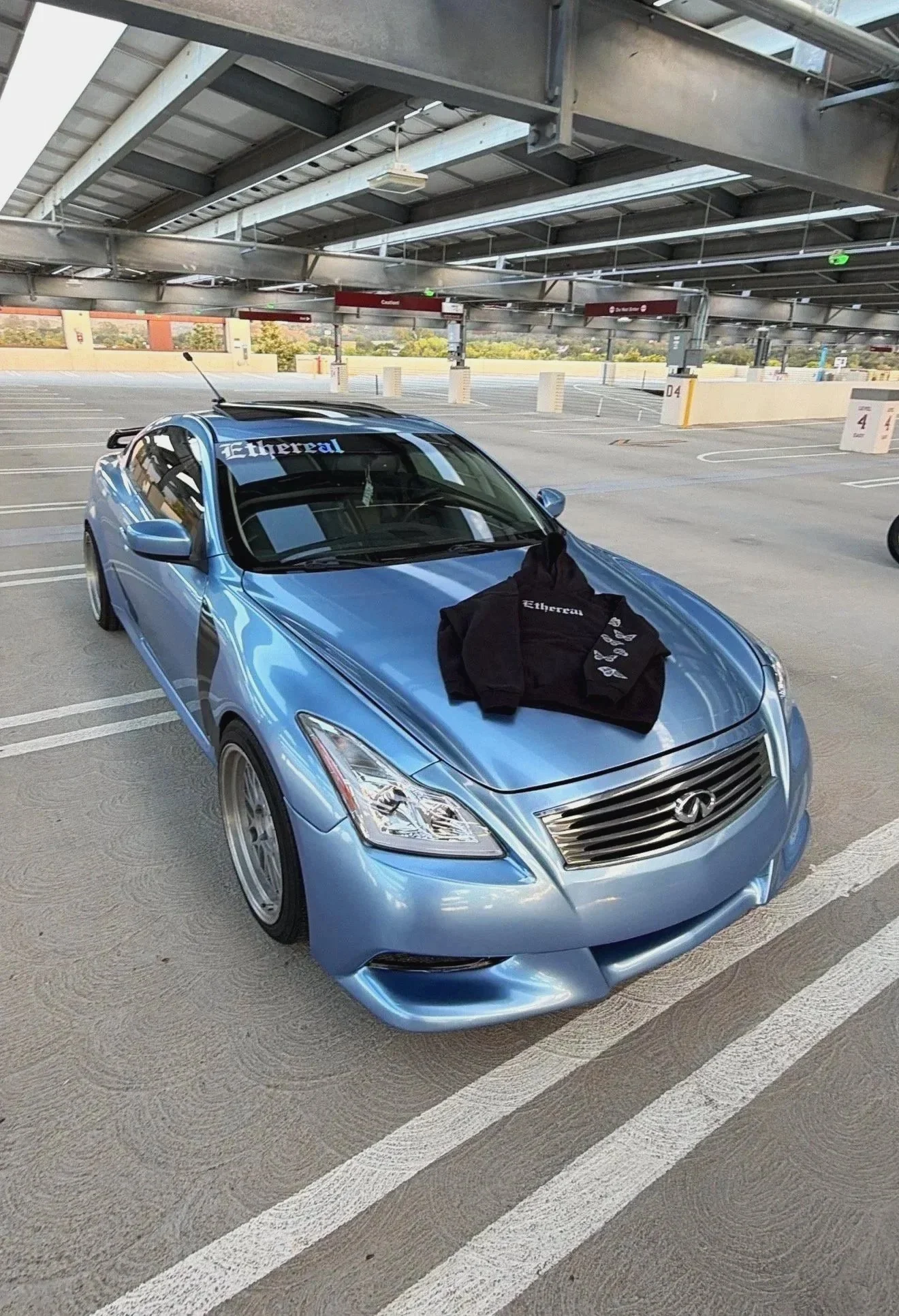A blue Infiniti car parked in an empty parking garage with a black hoodie draped over the hood, and a windshield decal that says 'Ethereal'.