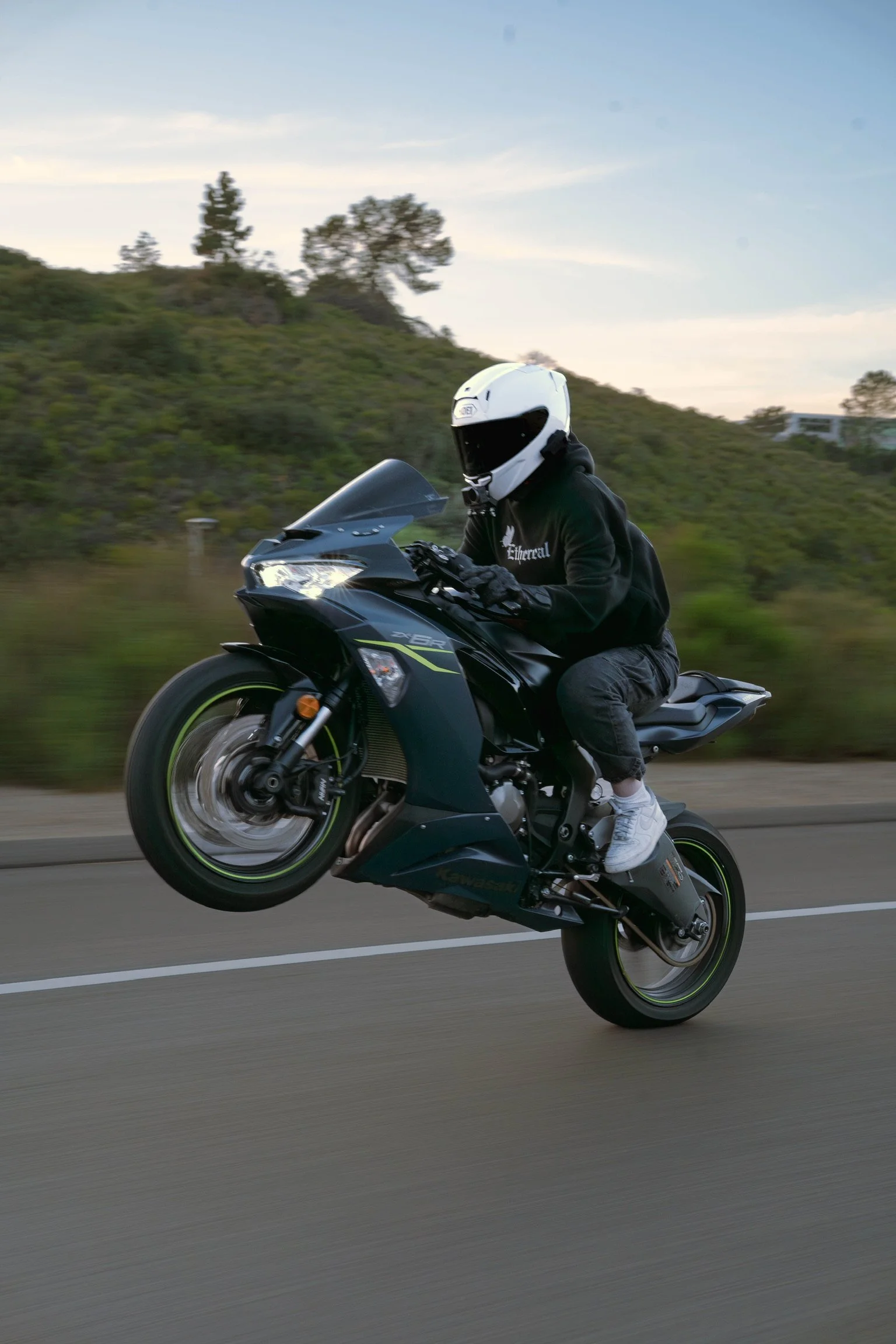 A person in a white helmet riding a black Kawasaki motorcycle on a road with greenery and hills in the background during daytime.