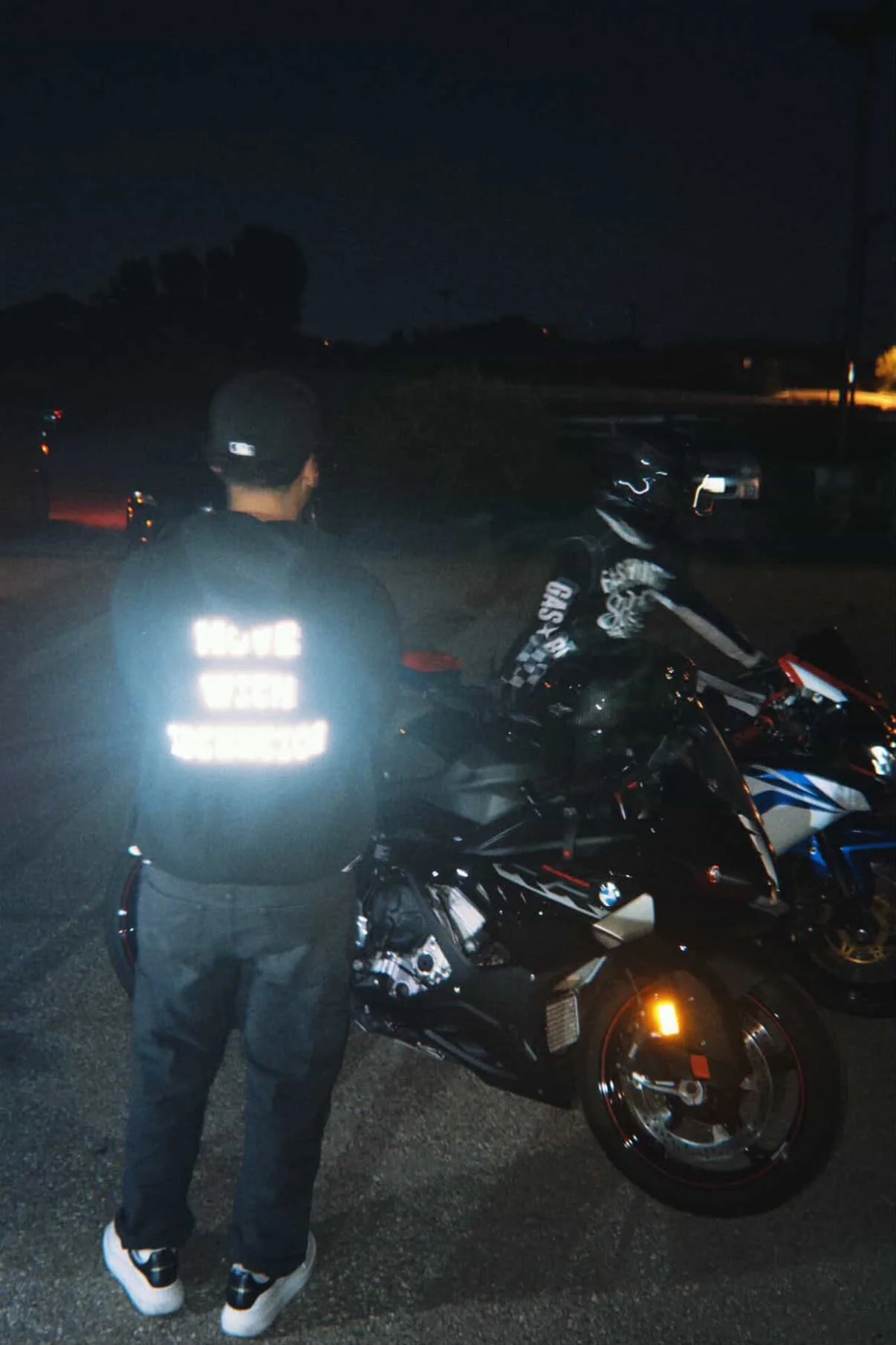 A person in a reflective vest standing next to a black motorcycle at night in a parking lot.
