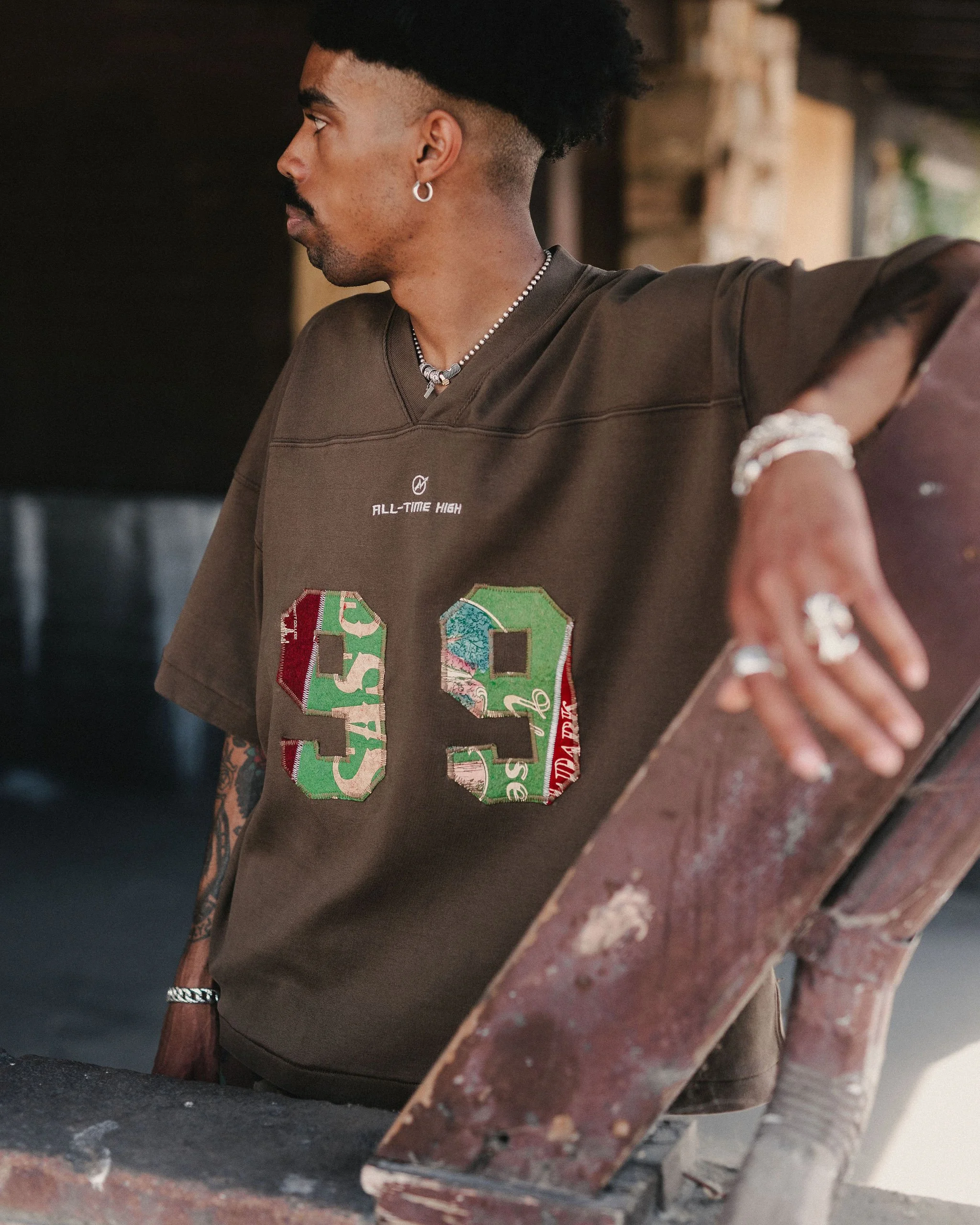 A man with a tattooed arm and hanging earrings, wearing a brown t-shirt with colorful embroidered letters, is leaning against a rusted metal railing in an industrial or rustic setting with stone and wood elements.