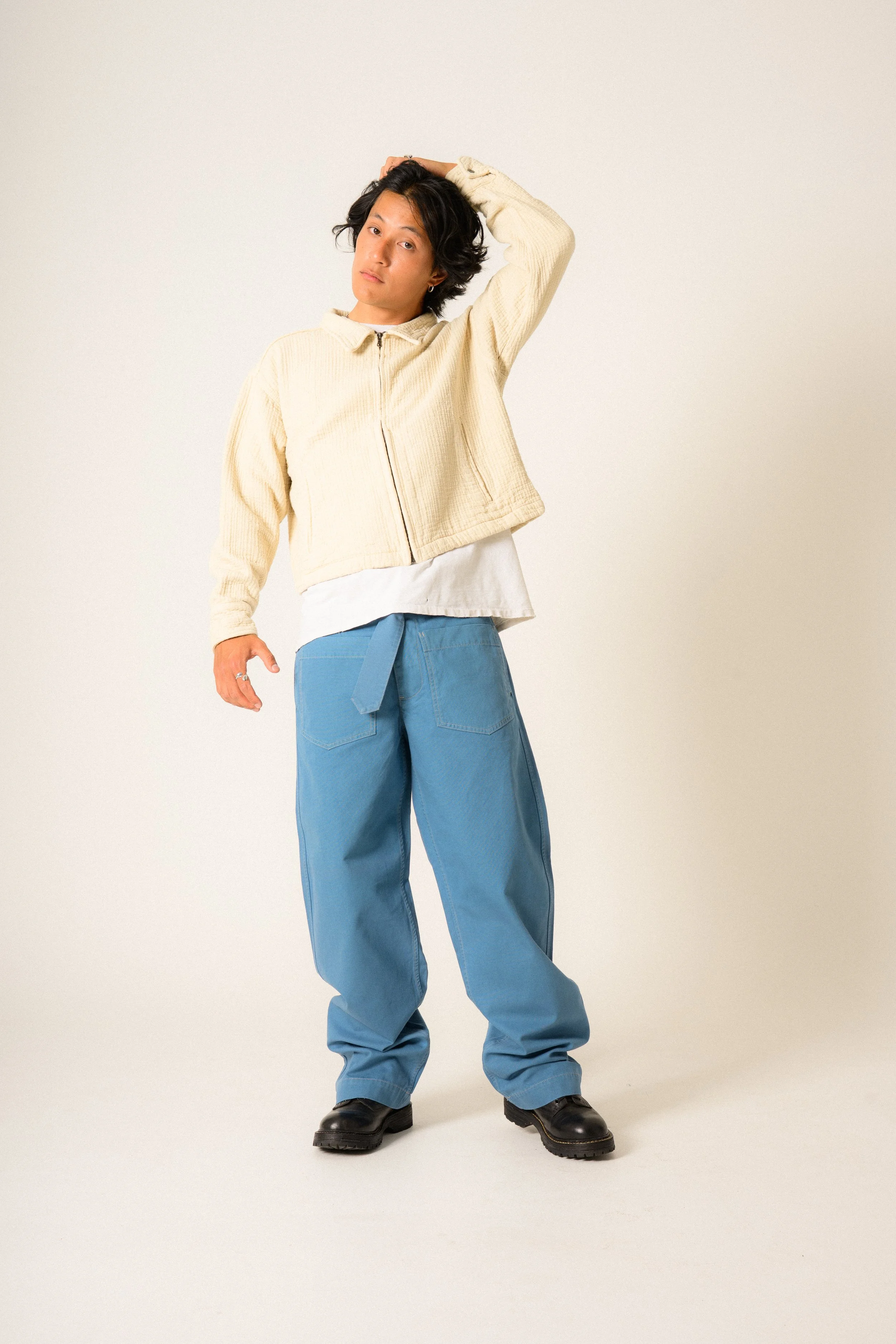 Man with medium-length black hair standing against a plain light background, wearing a beige zip-up jacket, white shirt, baggy blue jeans, and black boots, posing with one arm raised and hand on their head.