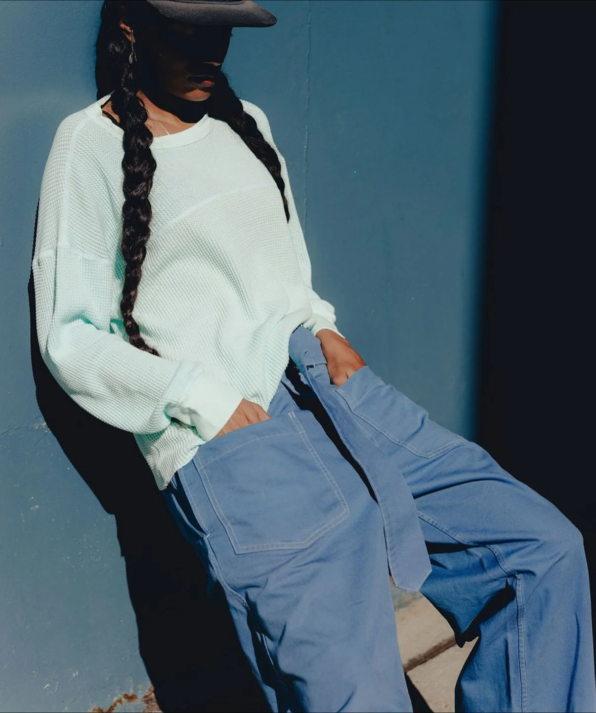 A woman with long braided hair wearing a black cap, white long-sleeve shirt, and blue cargo pants, leaning against a blue wall with hands in her pockets, looking down.