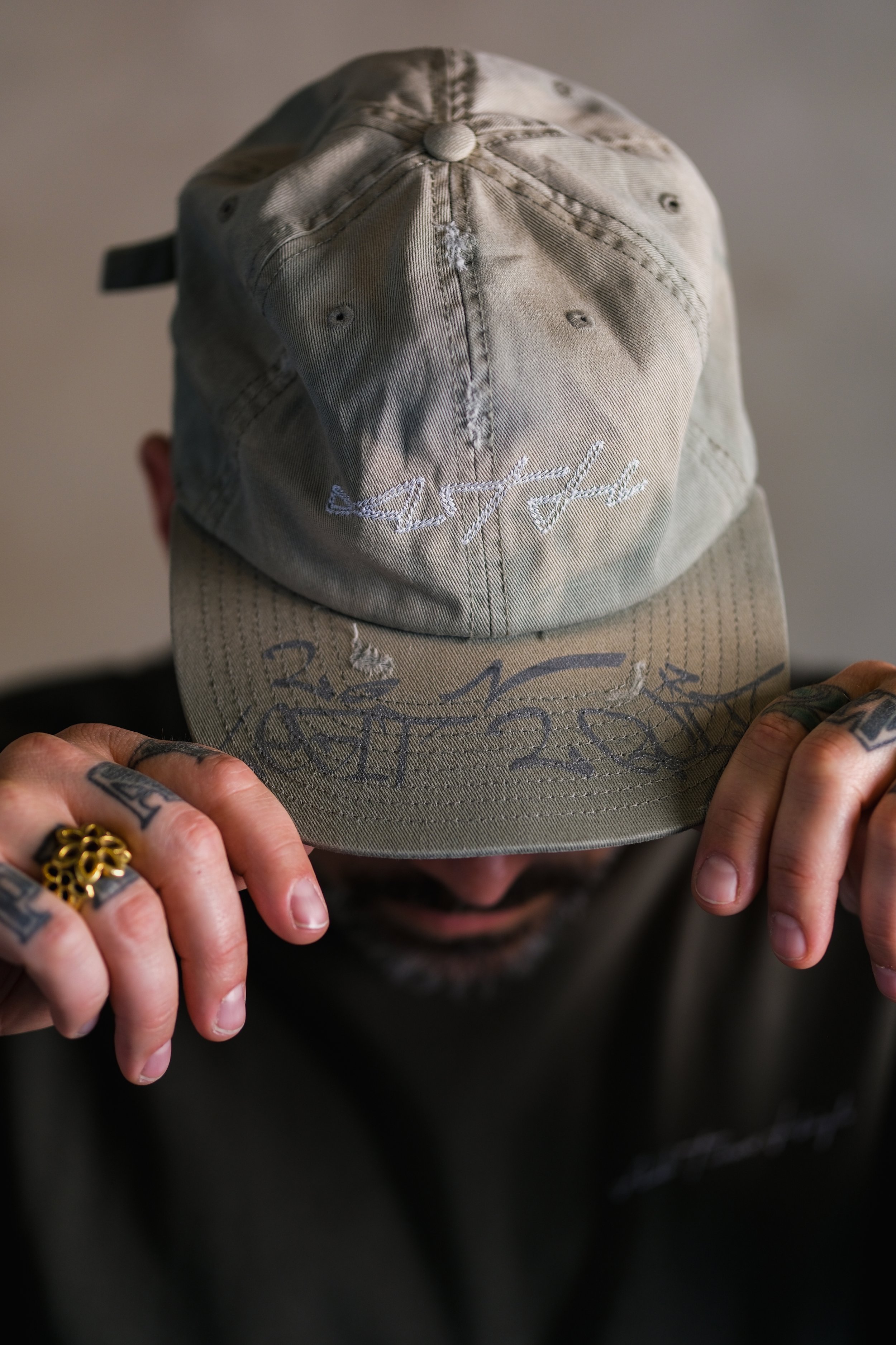 Close-up of a person wearing a beige cap with embroidered white text on the front, holding the brim with both hands, showing tattoos and multiple rings.