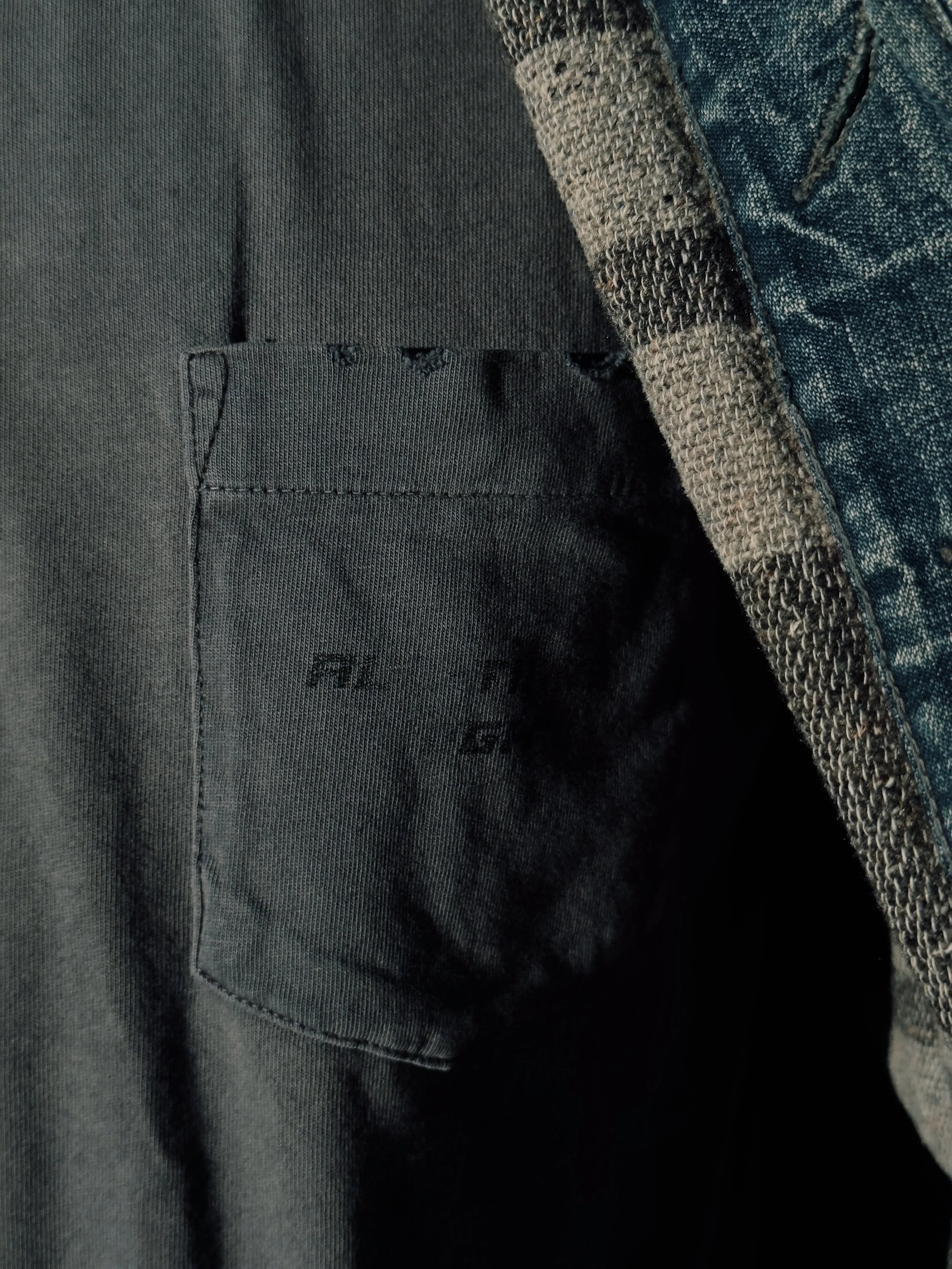 Close-up of a black pocket on a dark gray or black fabric garment, with a layered denim or textured fabric edge on the right side.