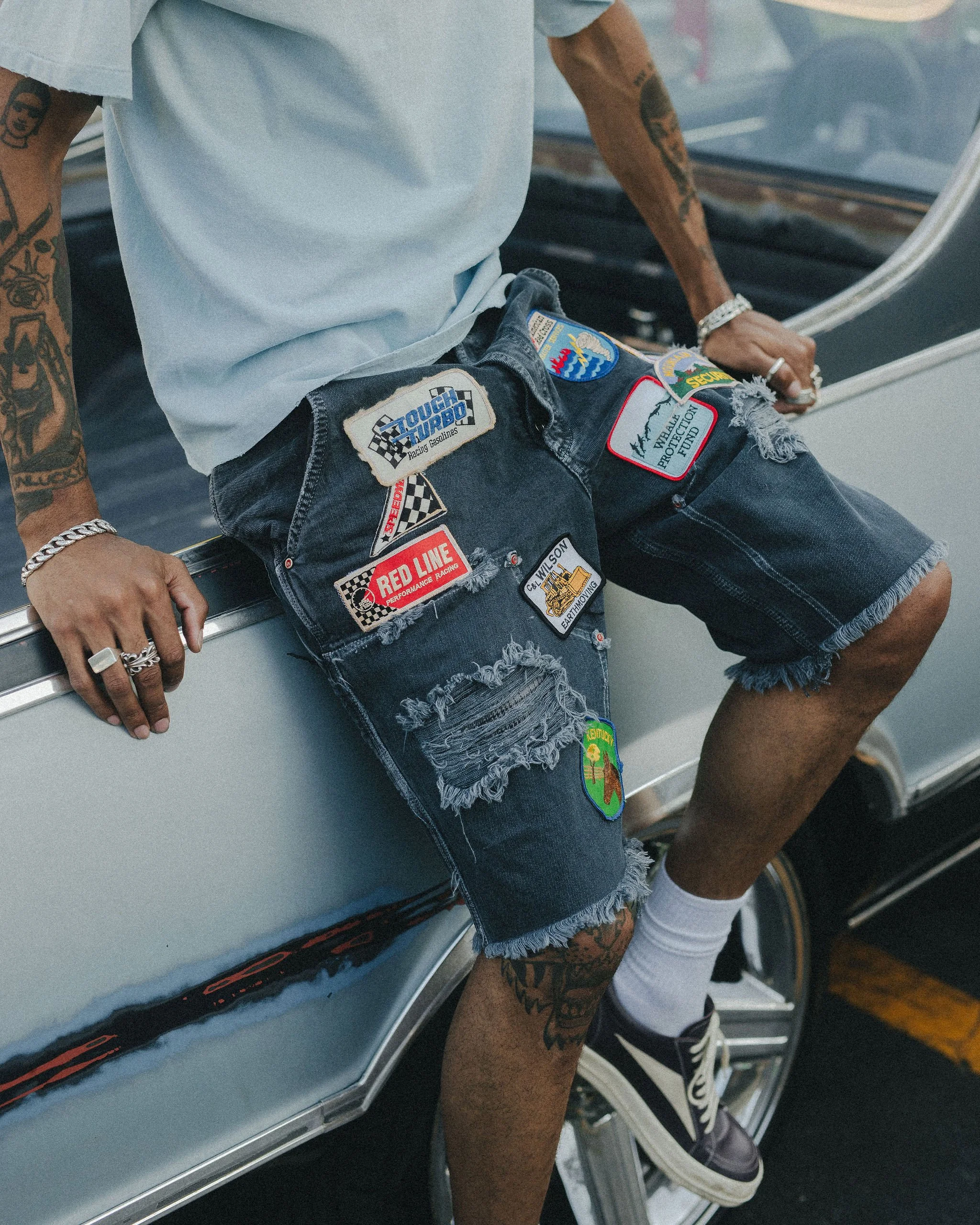 Close-up of a person wearing distressed denim shorts with various patches, including racing and adventure themes, sitting on a vintage car. The person has tattoos on their arms and hands, is wearing rings, bracelets, and sneakers, with a visible bracelet on the right wrist and hand resting on the car.