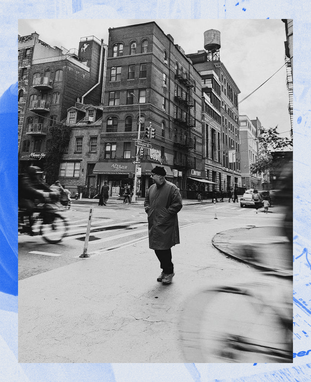 Black and white photo of a man walking on a city sidewalk at an intersection, surrounded by tall buildings, with some pedestrians and cyclists in the background.