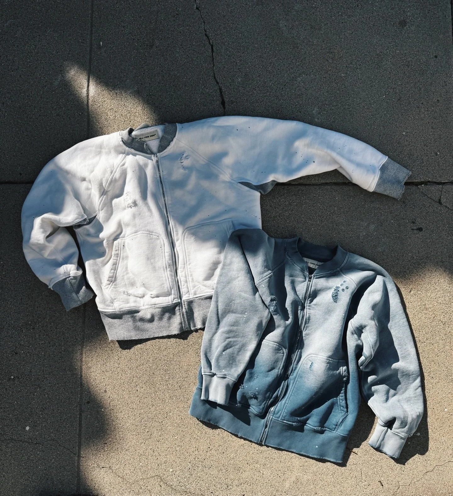 Two zip-up hooded sweatshirts, one white with grey cuffs and the other grey with distressed details, lying on a concrete sidewalk.