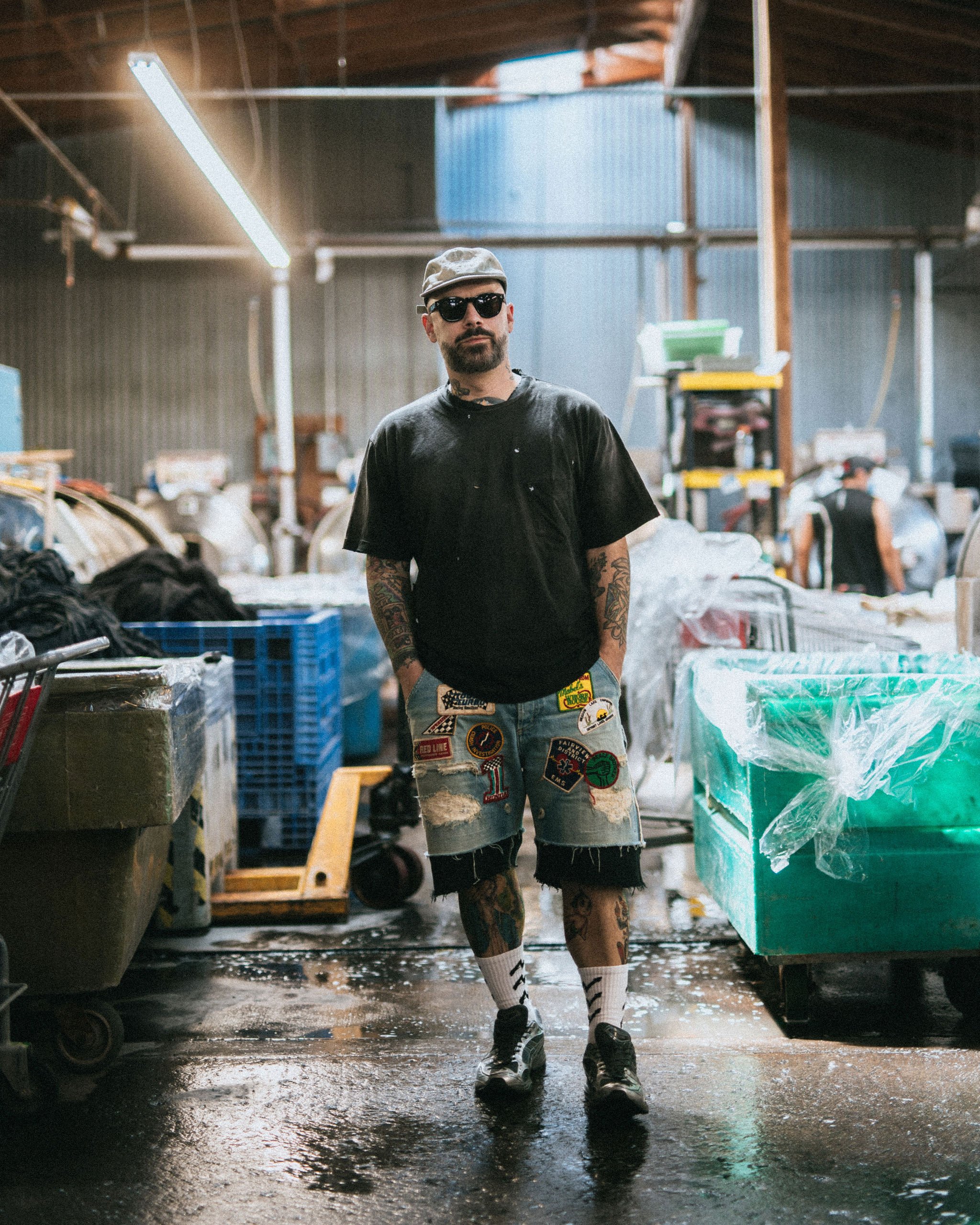 A man standing in a warehouse or industrial space with a wet and muddy floor, wearing sunglasses, a gray cap, a black t-shirt with paint splatters, and patched denim shorts with various patches. There are shelves, carts, and boxes around him.