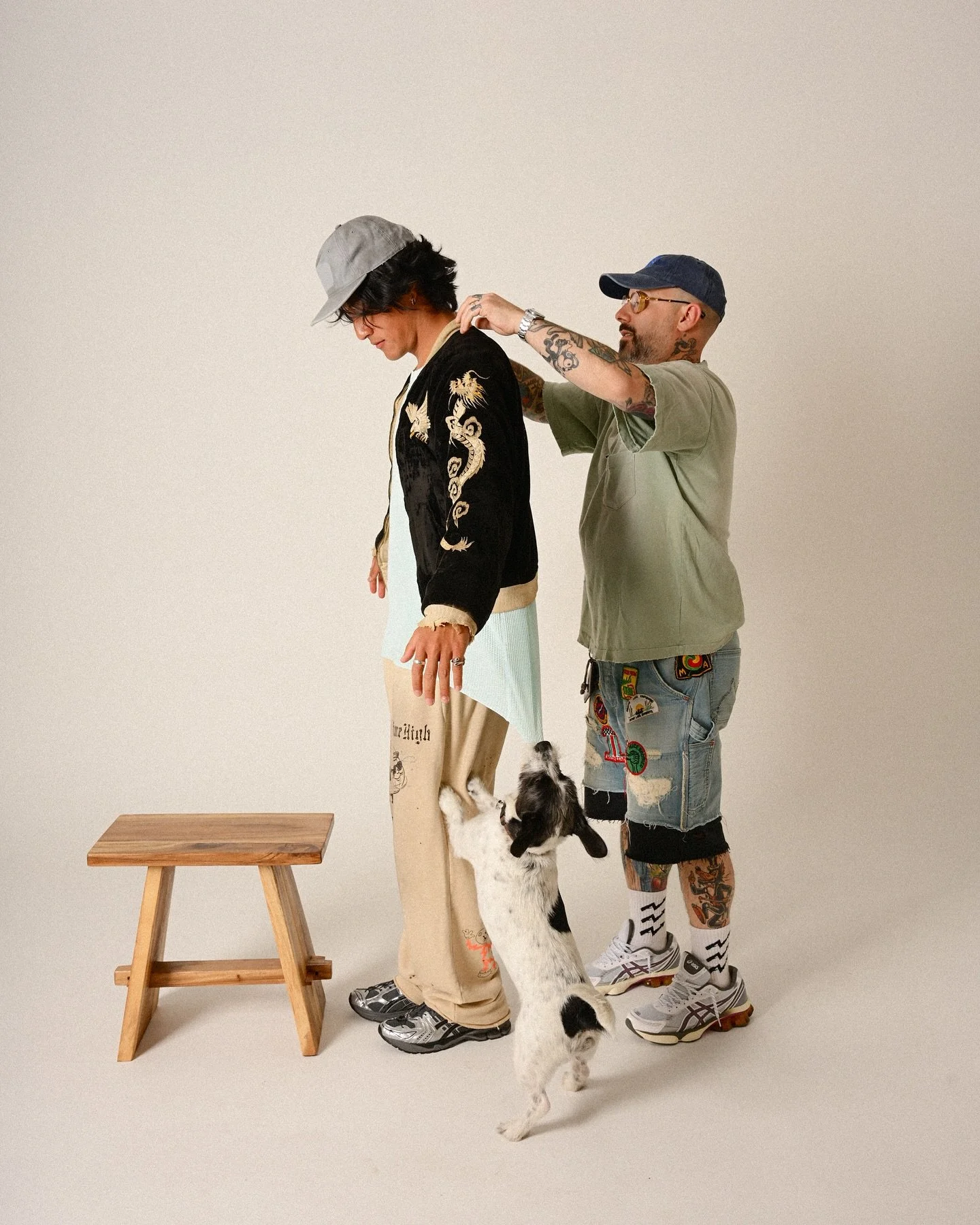 Two men and a dog in a studio with a plain background, one man is adjusting the other's collar, a small wooden stool is on the left.
