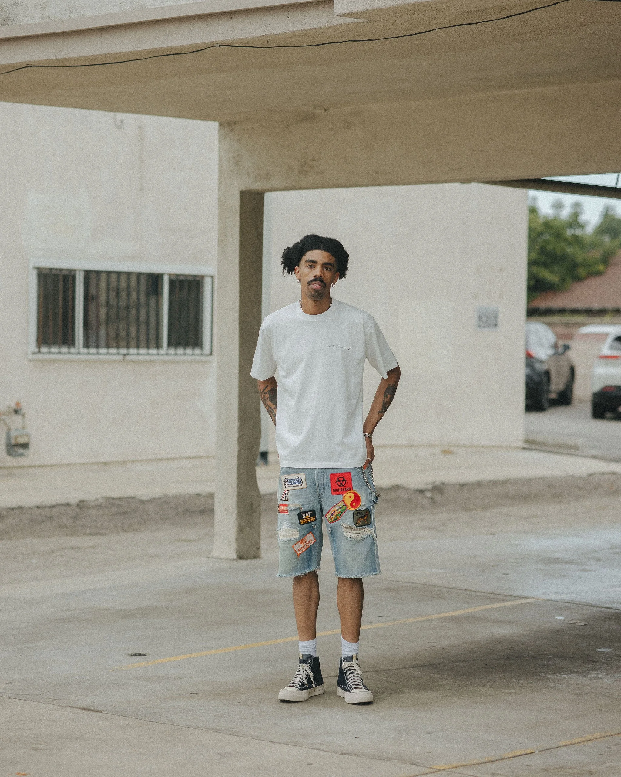 A man standing outside in a parking lot under a concrete structure, wearing a white oversized t-shirt, ripped denim shorts with patches, black and white sneakers, and socks, with tattoos on his arms and tattoos on his arms.