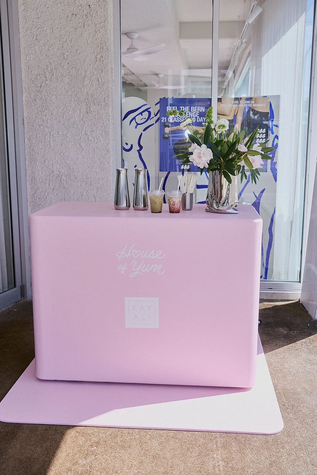 Pink table with a sign that says 'House of Yum' and 'Kay Ali'; a flower arrangement with white flowers and green leaves in a silver vase; metal shakers and cups with straws are placed on the table; in the background, a glass window with posters and a