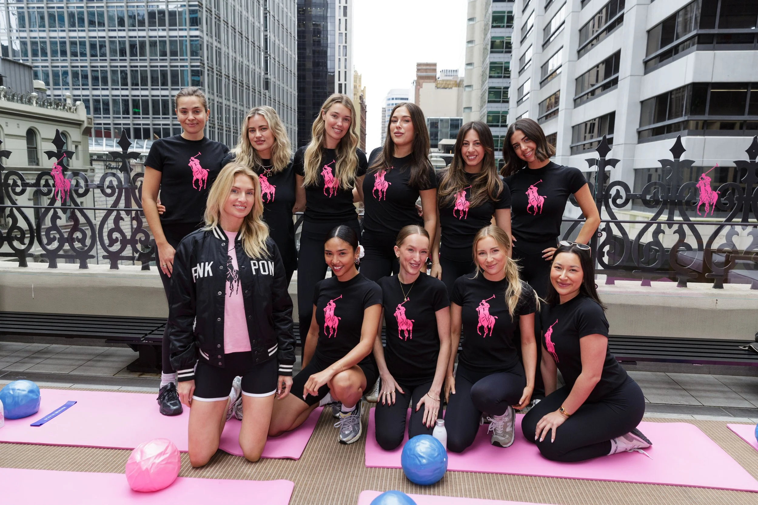 Group of women wearing black t-shirts with pink polo horse logo, posing outdoors in a cityscape with tall buildings, on with pink and beige mats, surrounded by colorful balloons.