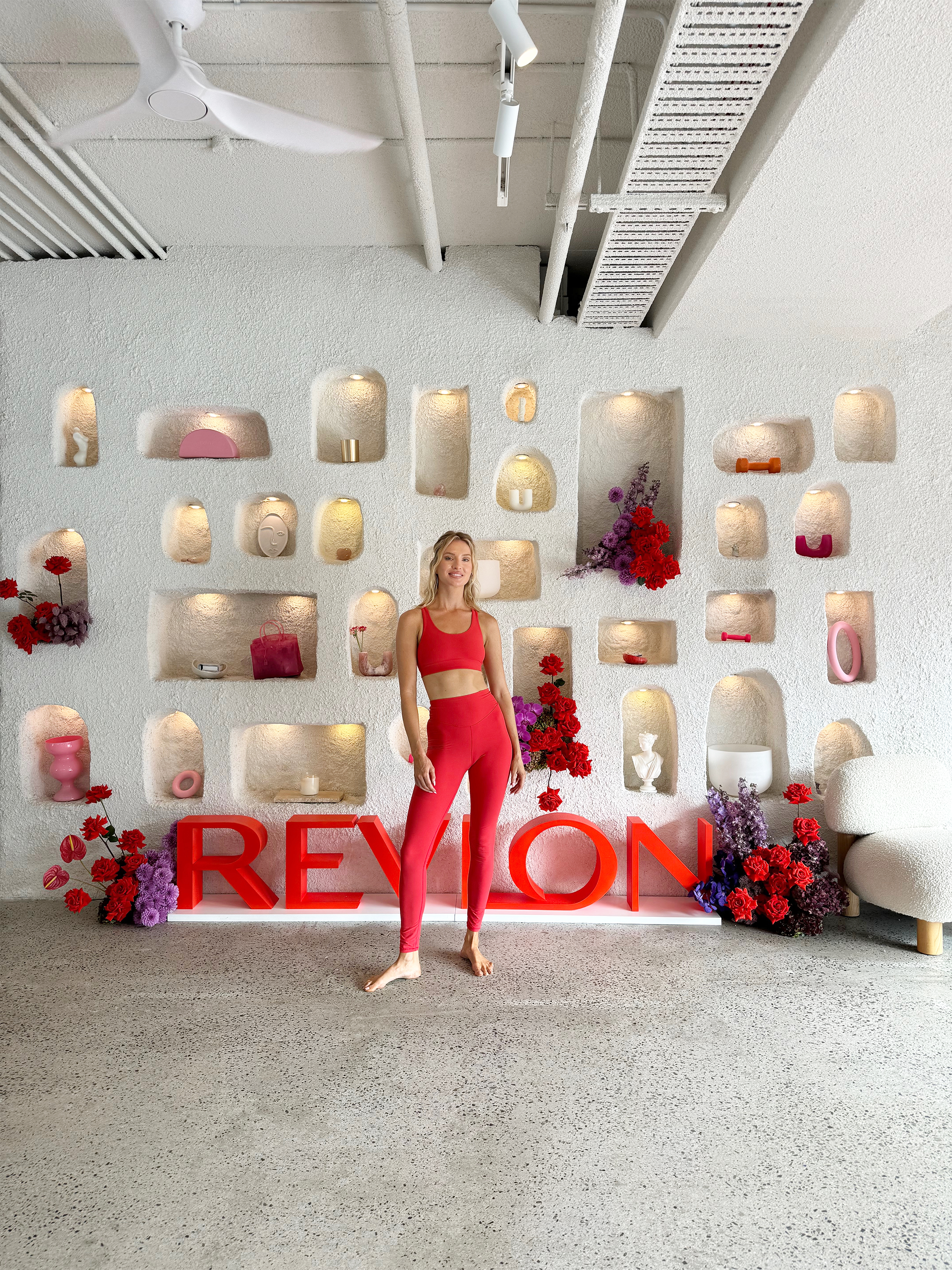 A woman in red workout clothing standing in front of a decorated wall with the word "REVLON" displayed in large red letters, surrounded by flowers and decorative objects.