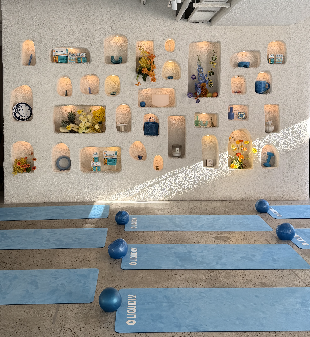 Yoga studio with blue mats and exercise balls in front of a decorative wall with various niches holding vases, flowers, candles, and decorative items, illuminated with soft lighting.