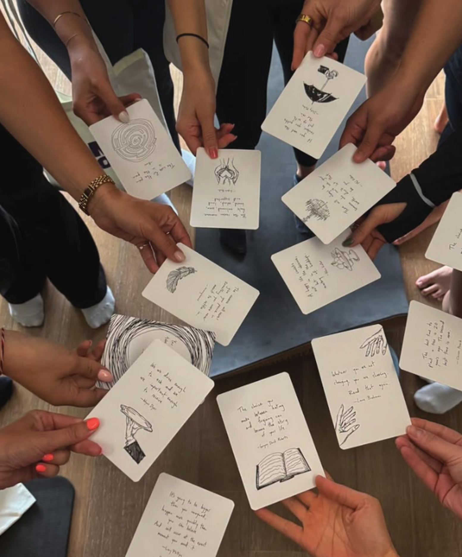 group of people holding playing cards with handwritten notes and illustrations, gathered around a wooden table.
