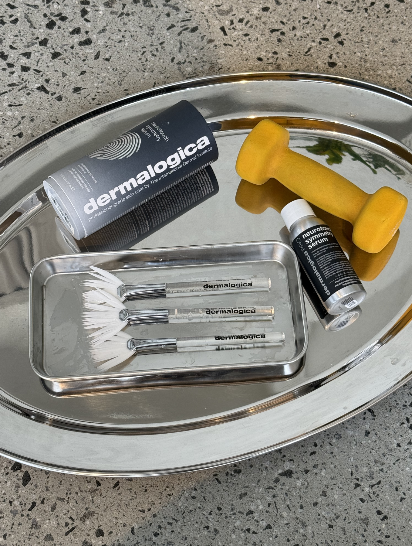 A silver tray with skincare and beauty tools, including a tube of Dermalogica skincare, a small roller bottle of Neurotoucher serum, a yellow massage roller, and a set of four white facial cleansing brushes with clear handles labeled 'dermalogica' on