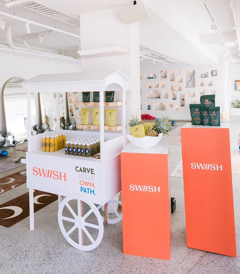 A white cart with products and the signs 'SWISH' and 'Carve your own path' on it. The cart displays bottles of yellow and dark liquids and is positioned next to two orange pedestals with products in green packaging. The background features a decorate