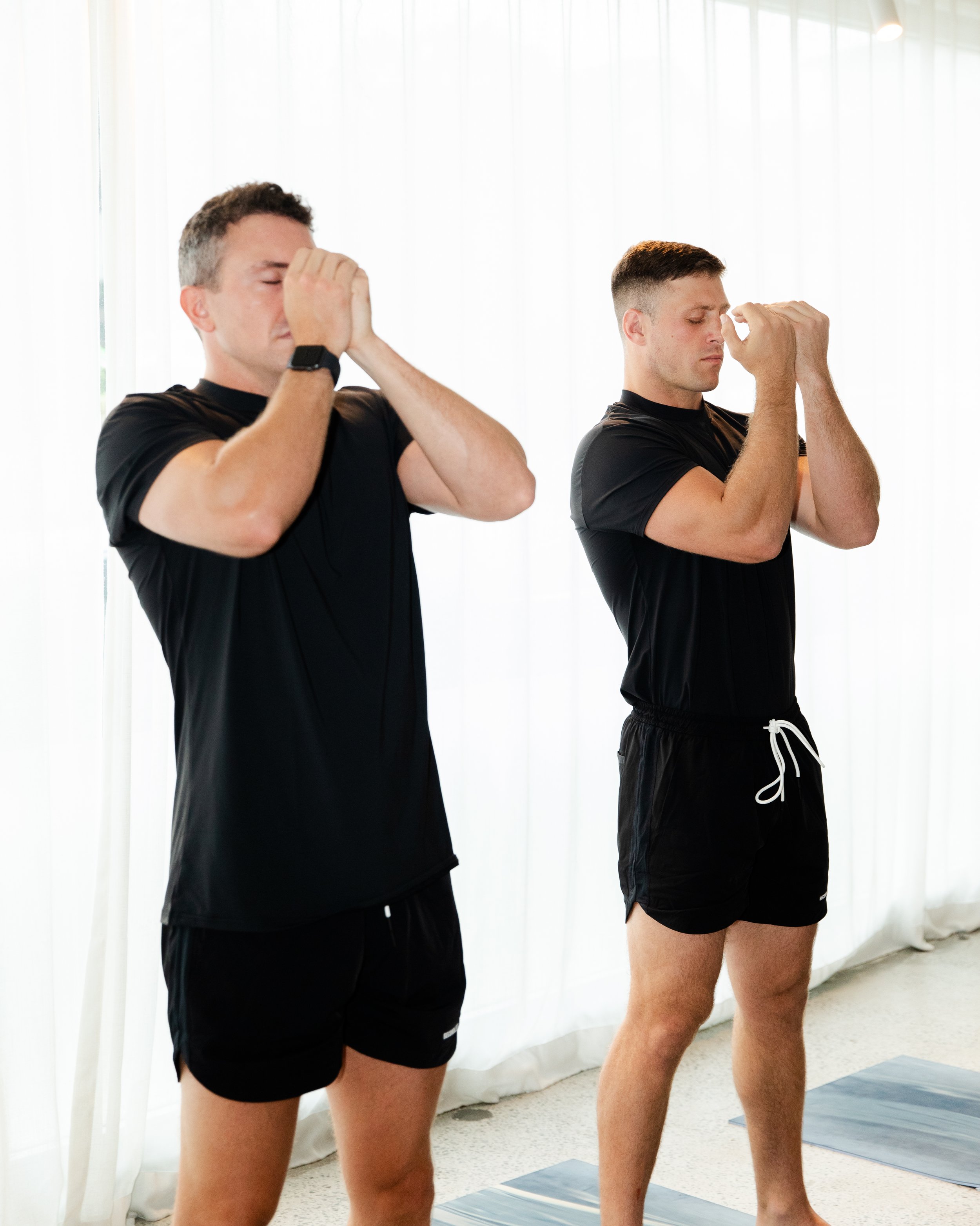 Two men standing with eyes closed in a meditation or prayer pose, with hands pressed together in front of their faces, indoors in front of a window with sheer curtains, wearing black athletic clothing and shorts.