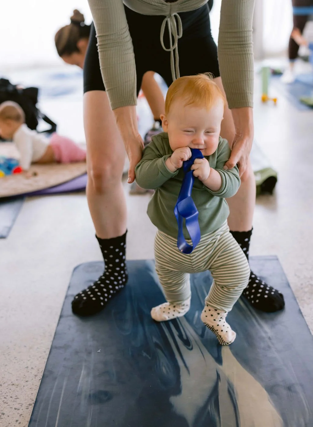 Baby learning to stand on a black mat with assistance from a woman, holding a blue ribbon in baby's mouth, with other children and toys in the background.