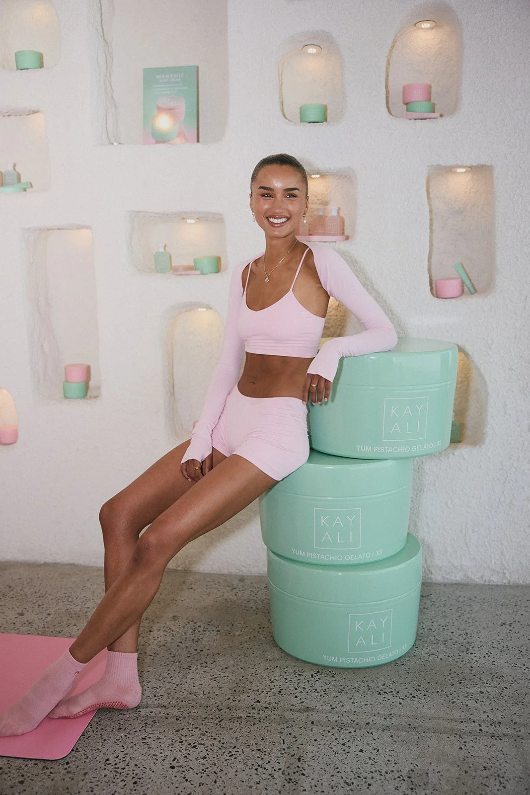 A woman in pink loungewear sitting on stacked pastel green containers labeled 'Kay Ali Yum Pistachio Gelato' in a minimalist, pastel-colored room with lit wall niches and decorative objects.