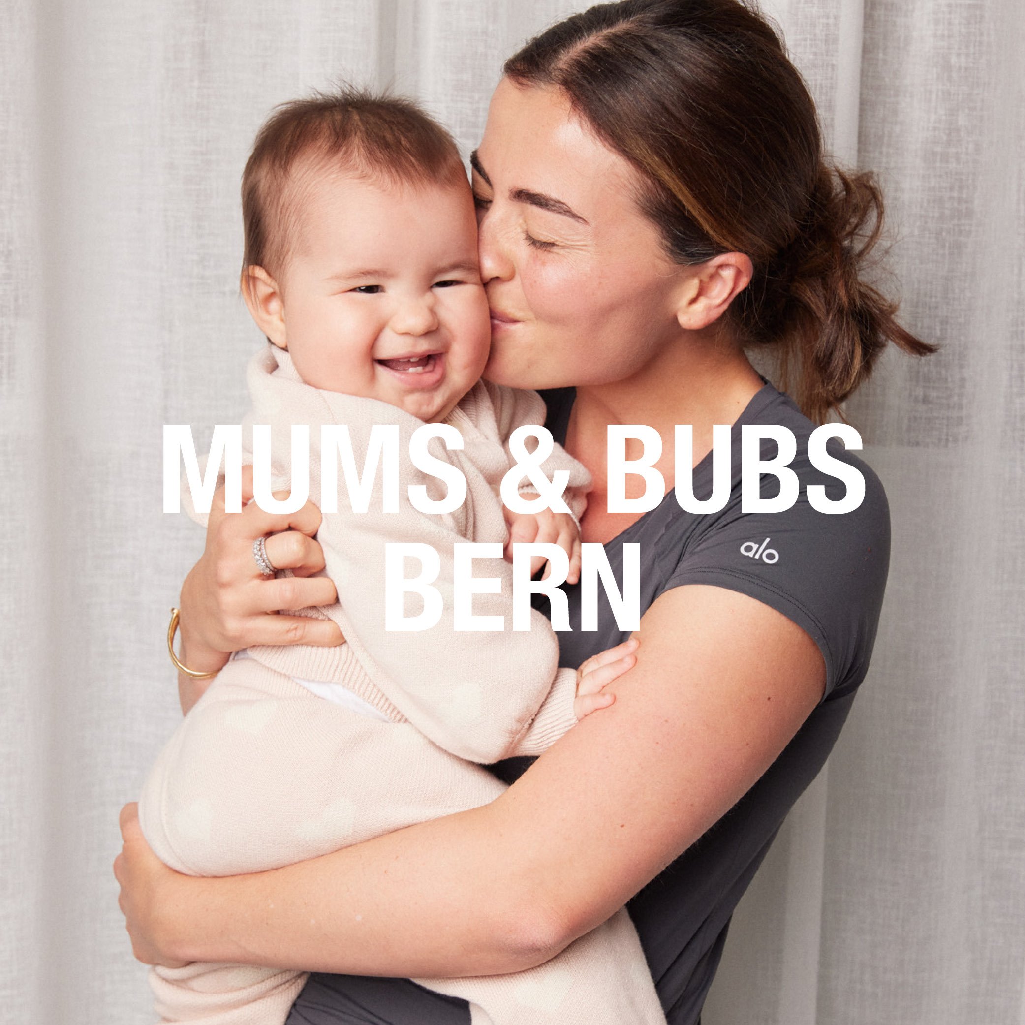A woman with brown hair and a gray shirt holding a smiling young child with short light brown hair and a beige sweater, hugging tightly in front of a plain light-colored curtain.
