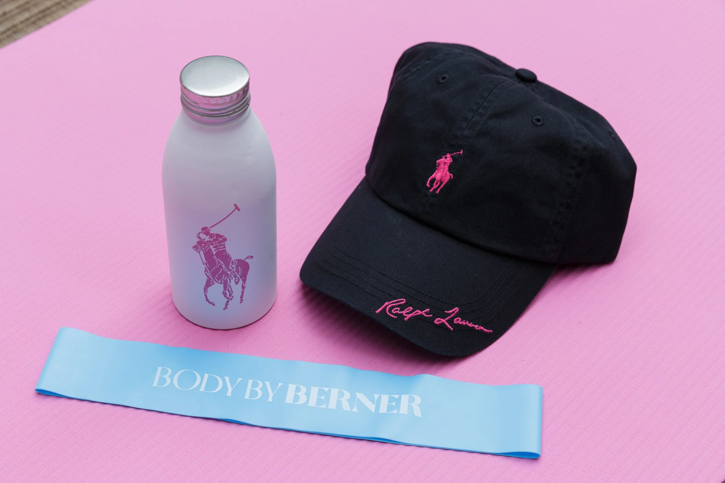 A black baseball cap with pink embroidered logo and pink signature, a white water bottle with pink logo, and a blue ribbon with white text that says "BODY BY BERNER" on a pink background.