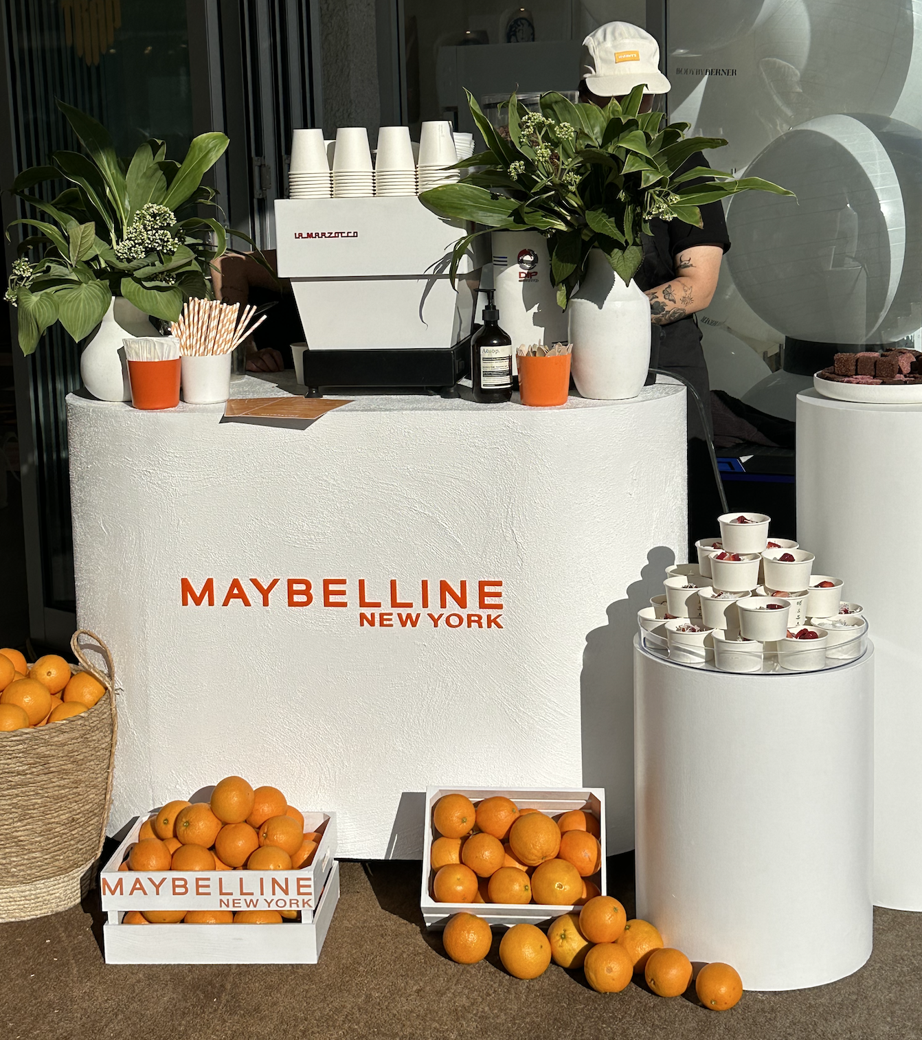 A Maybelline New York promotional display at a storefront featuring a white counter with orange text, baskets of oranges, several small cups of yogurt topped with berries, potted green plants, white cups stacked behind the counter, a spray bottle, an