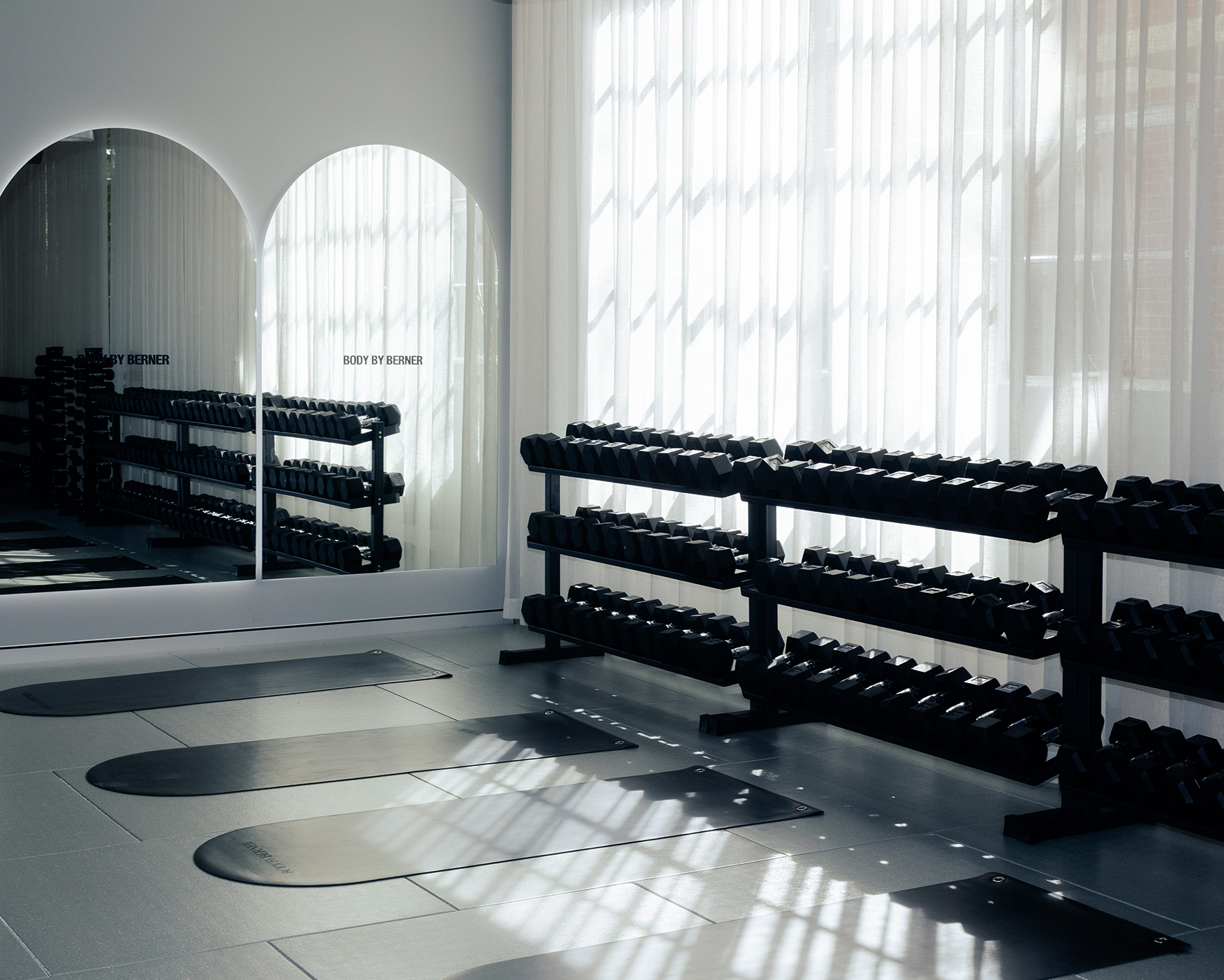 A gym with black dumbbell racks near a window with sheer curtains, sunlight casting shadows on the floor, and exercise mats placed in front of the mirrors.