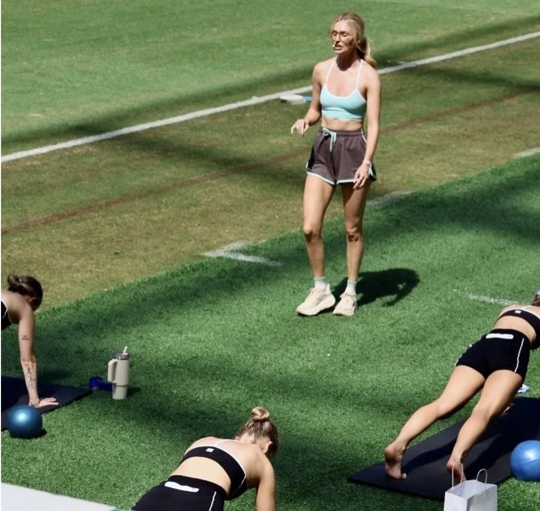 Fitness instructor standing outdoors while three women in workout clothes do push-ups on yoga mats.
