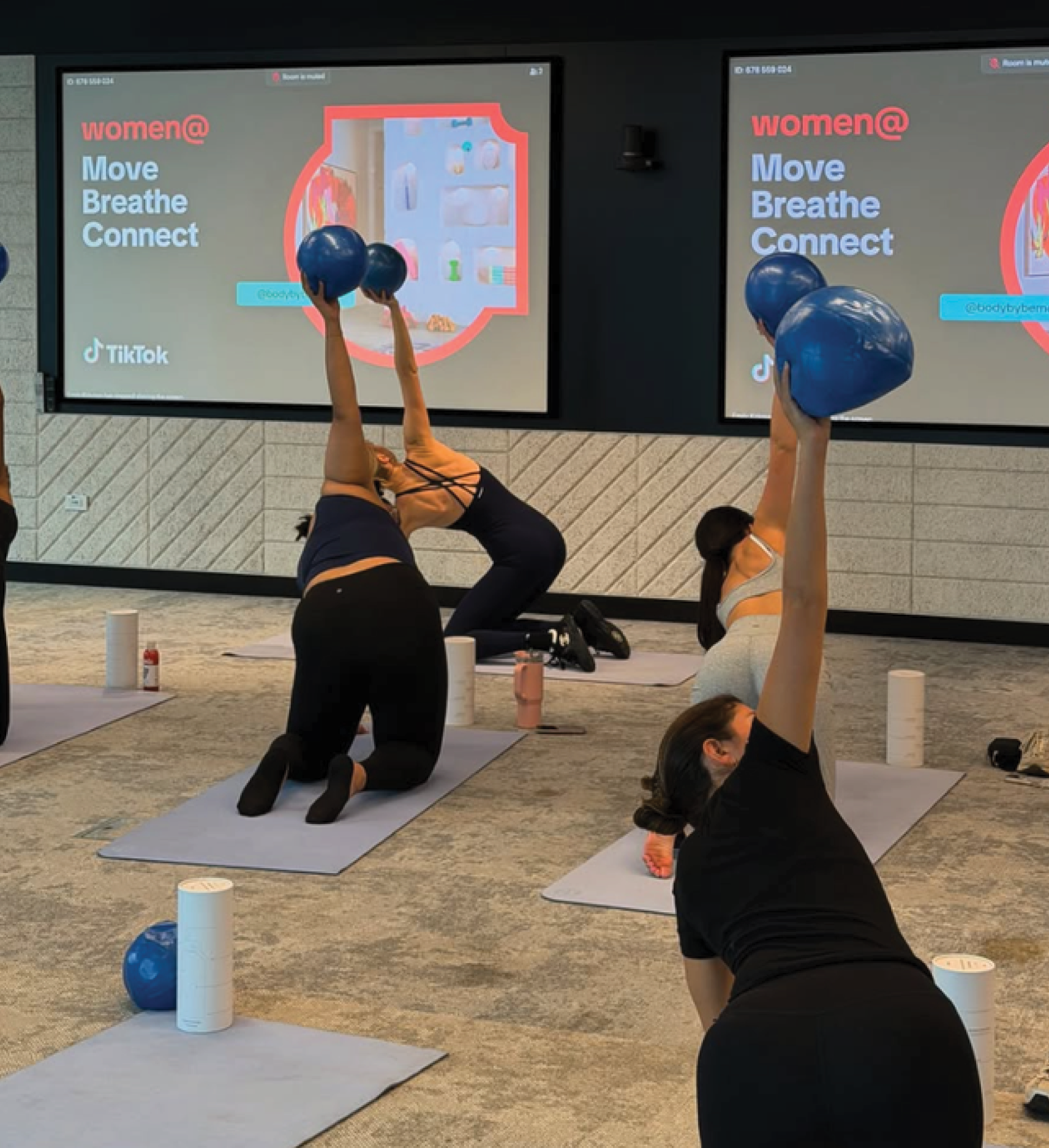 People participating in a yoga or fitness class, doing a stretching pose with one arm raised and holding a blue exercise ball, on gray mats in a room with large screens displaying the words 'women@', 'Move Breathe Connect', TikTok logo, and a website