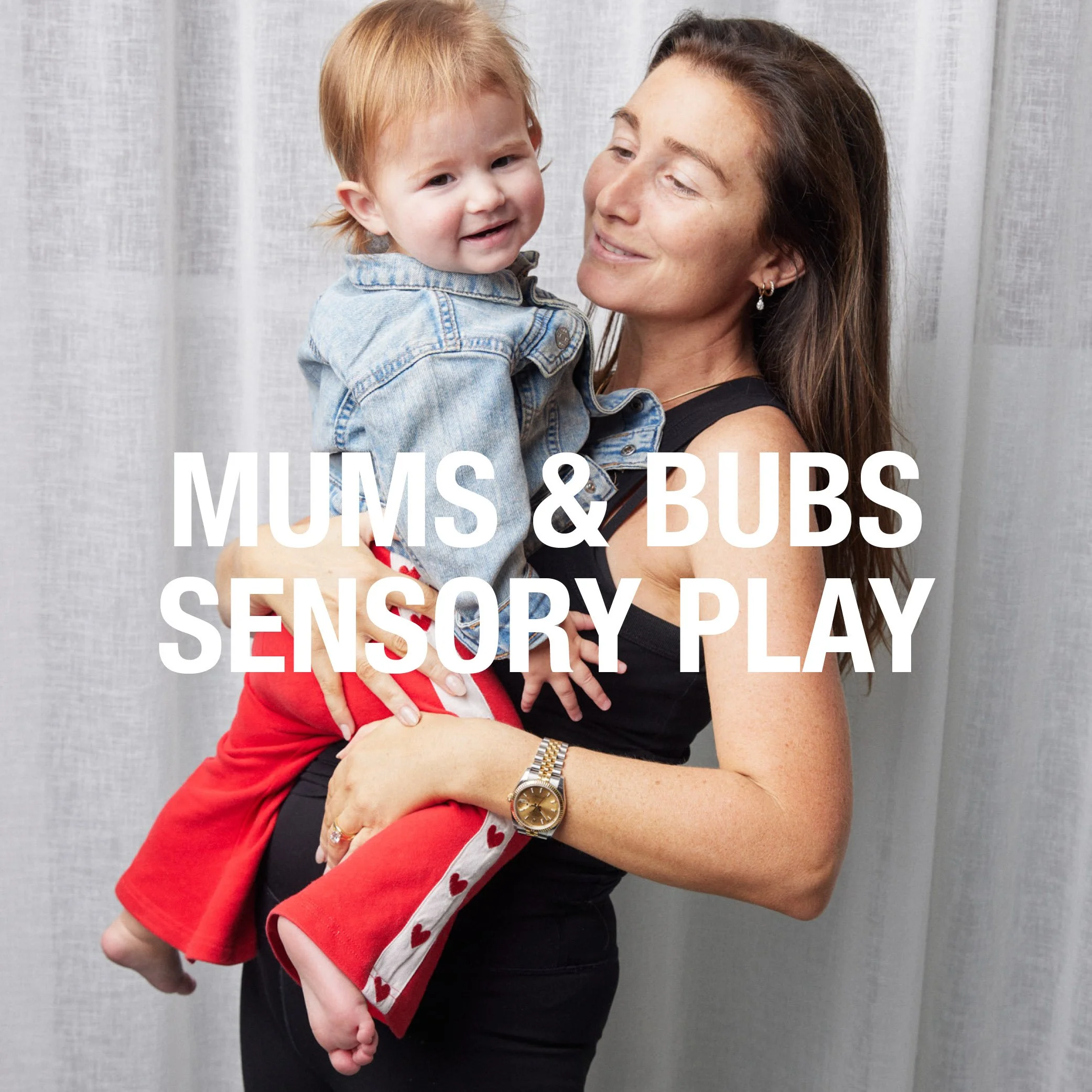 A woman holding a young boy with blonde hair, both smiling and looking at each other, against a light gray background. The boy is wearing a denim jacket and red pants with heart patterns.