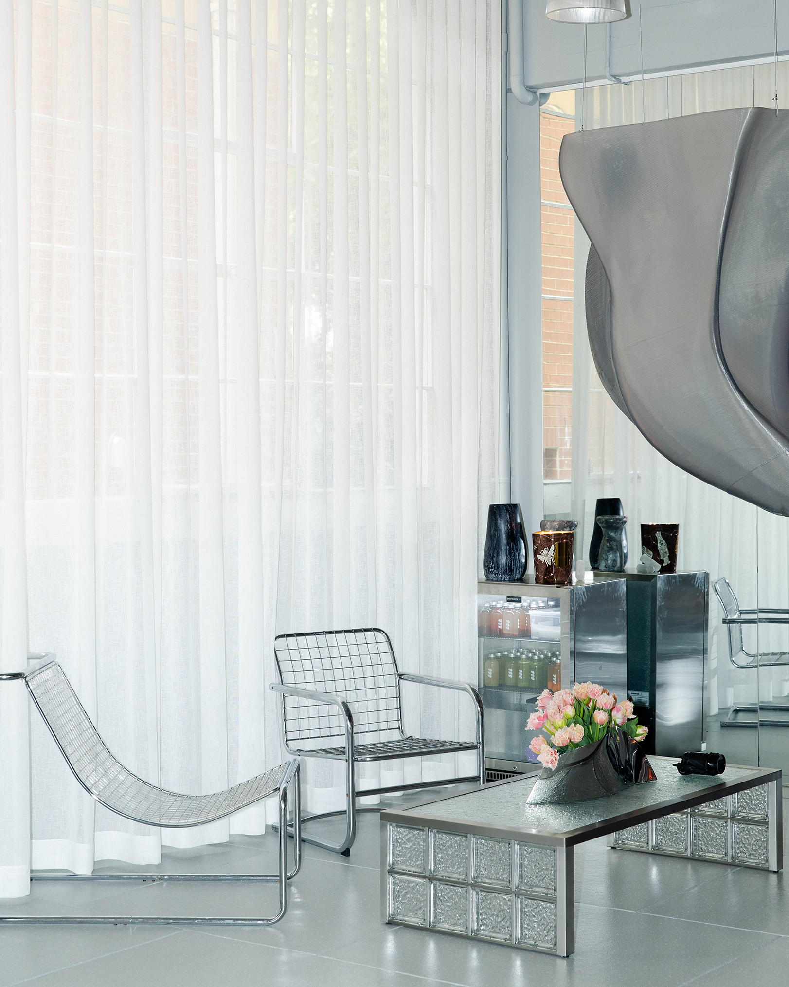 Modern lounge area with metallic chairs, a glass table with pink flowers, a small fridge with drinks, and large sheer curtains allowing natural light.
