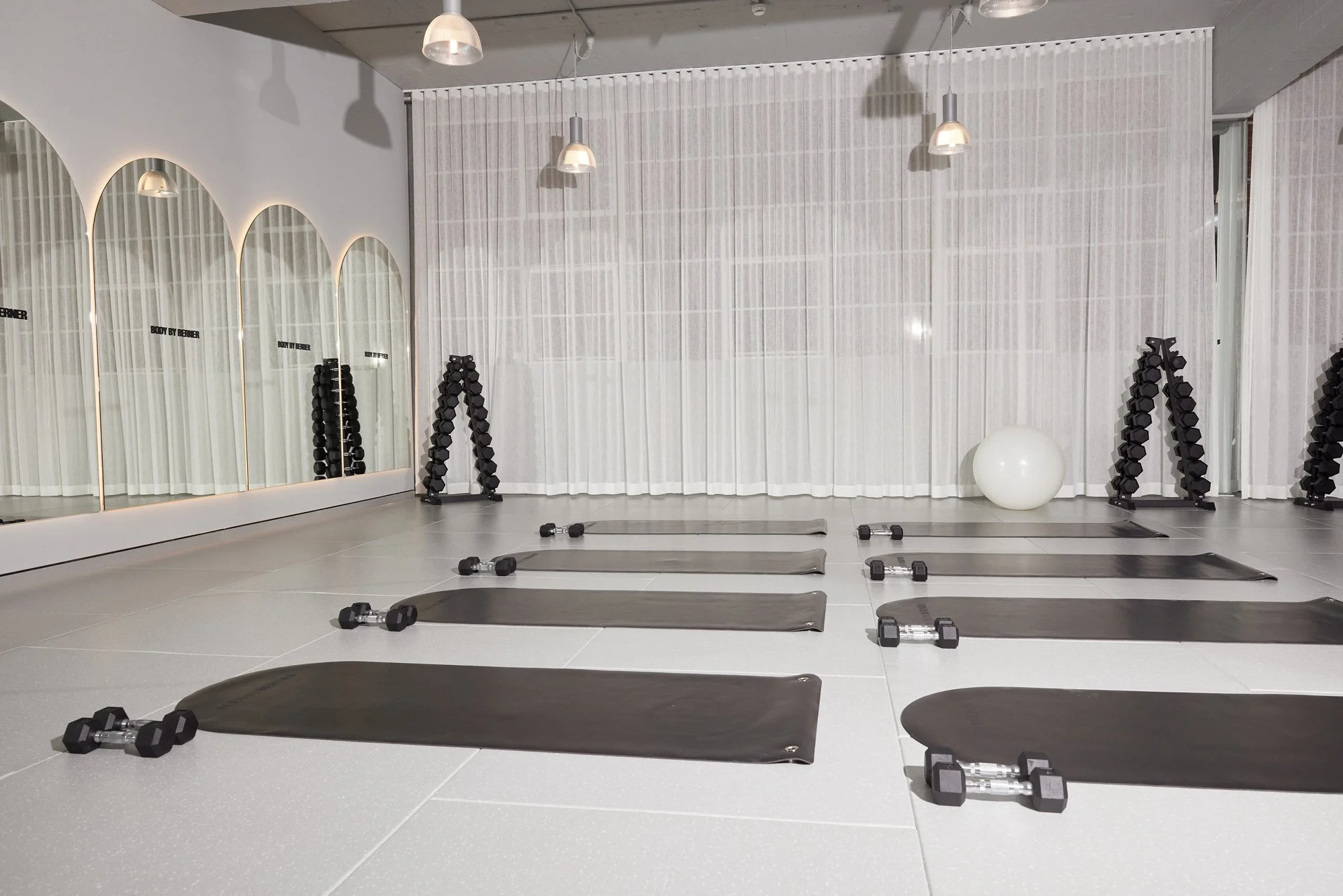 A modern fitness studio with four yoga mats, dumbbells, and exercise equipment, featuring three large mirrors and hanging lighting fixtures.
