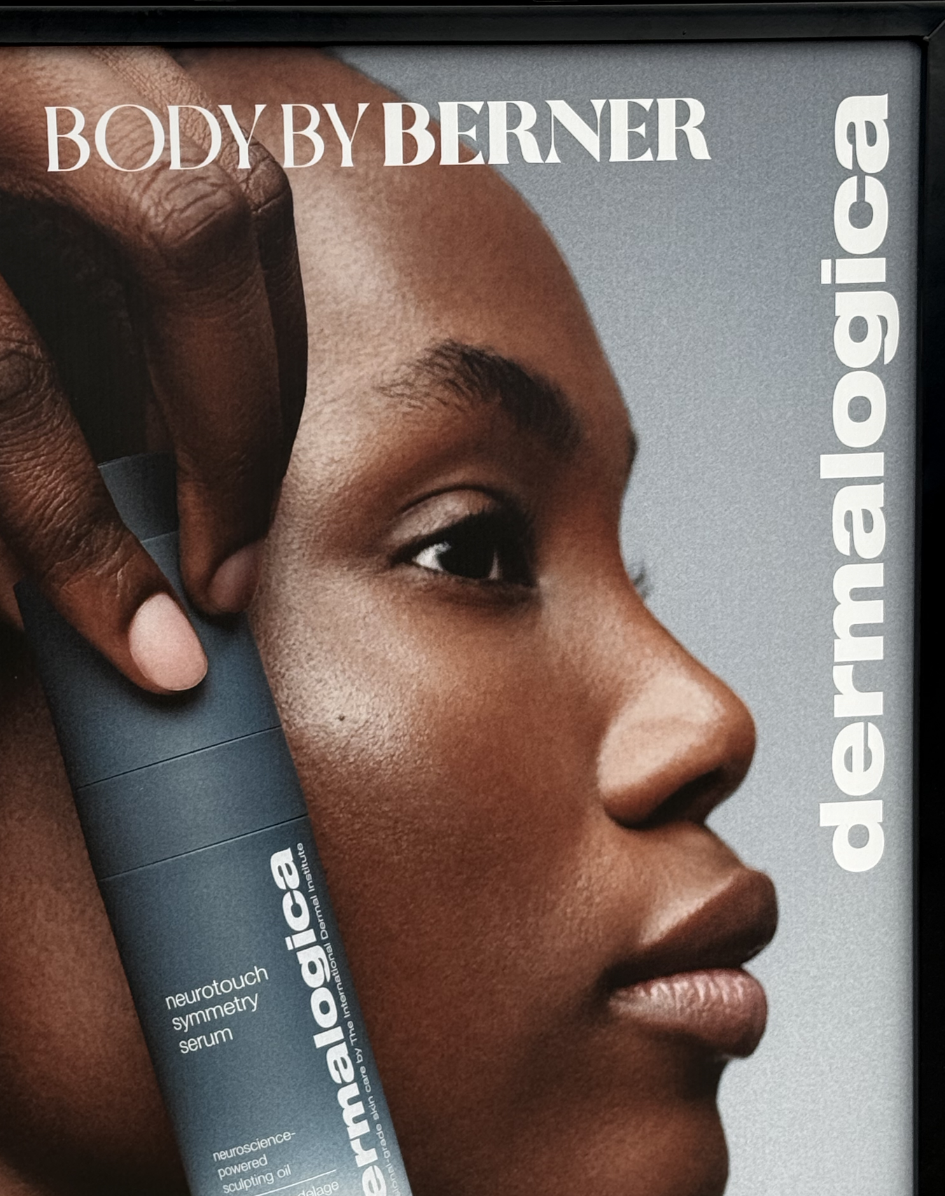Close-up of a woman's face with smooth skin, showing her eye, nose, and lips. A hand is holding a gray cylindrical container near her temple, which appears to be a skincare product. The text on the container reads "neurotouch symmetry serum" and it i
