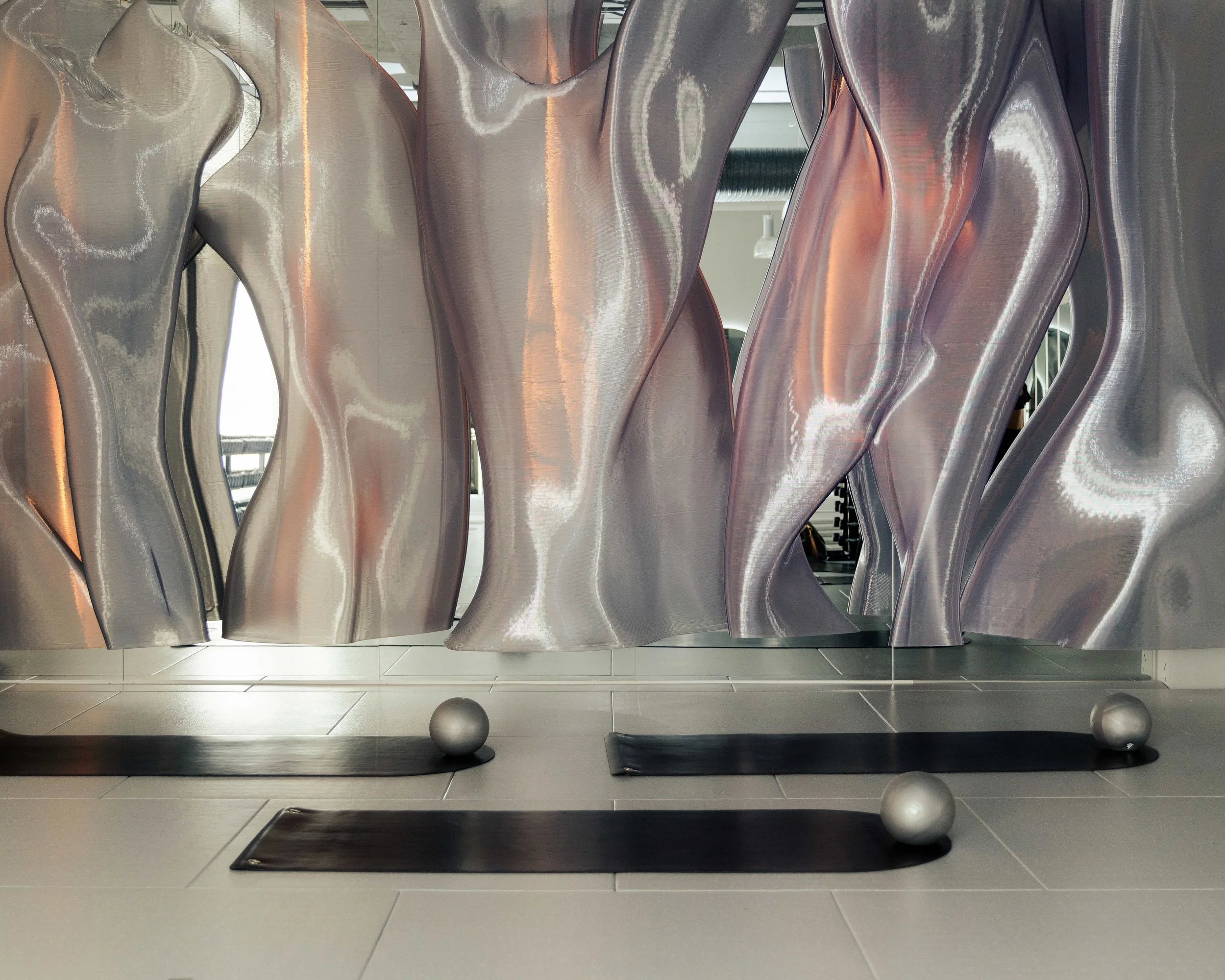 Metallic abstract sculpture with flowing, curvy shapes and reflective surfaces, displayed indoors on a tiled floor.