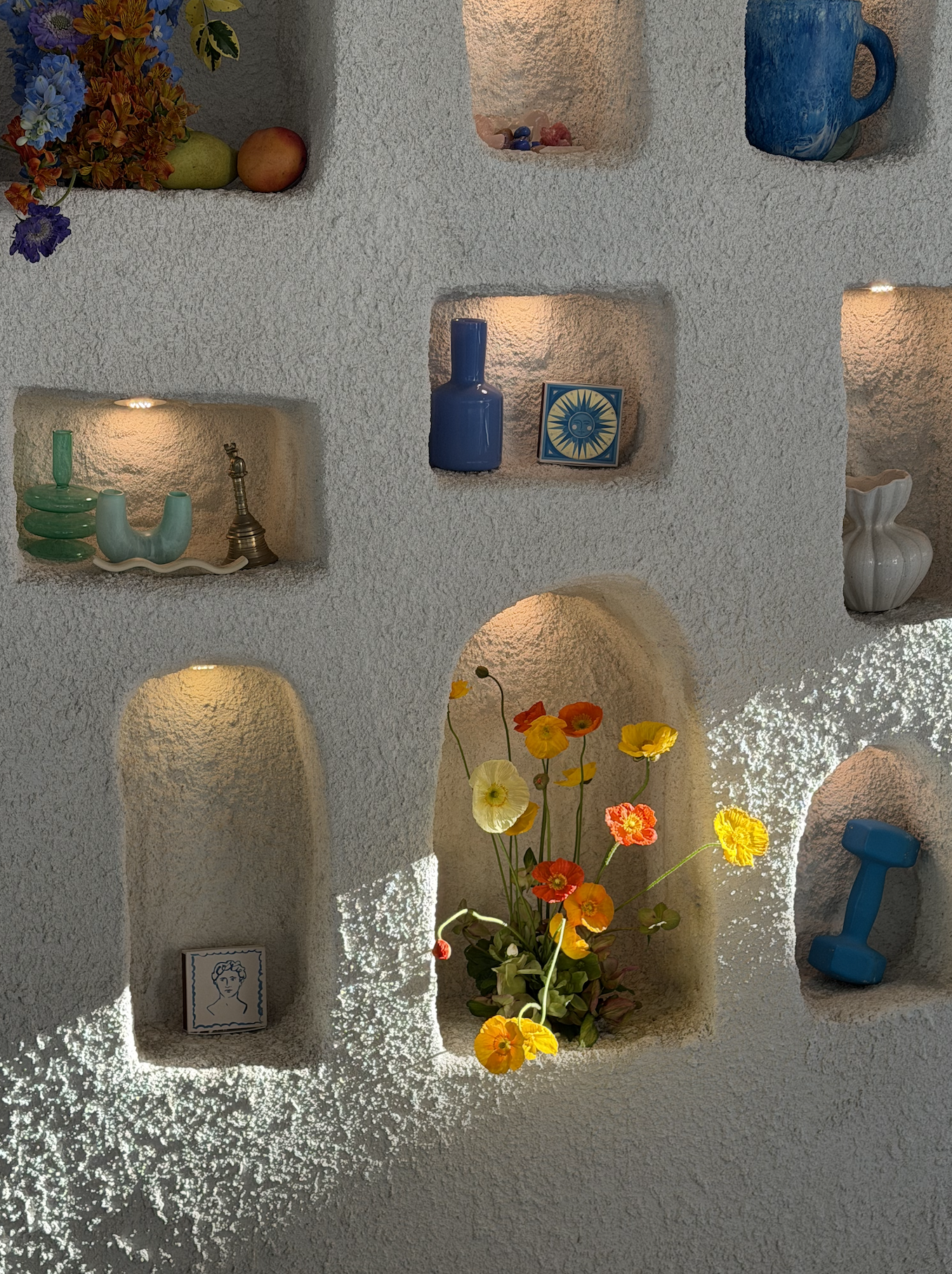 Decorative white plaster wall with built-in shelves, displaying colorful vases, a small artwork, flowers, and fruits, with sunlight illuminating the textured surface.