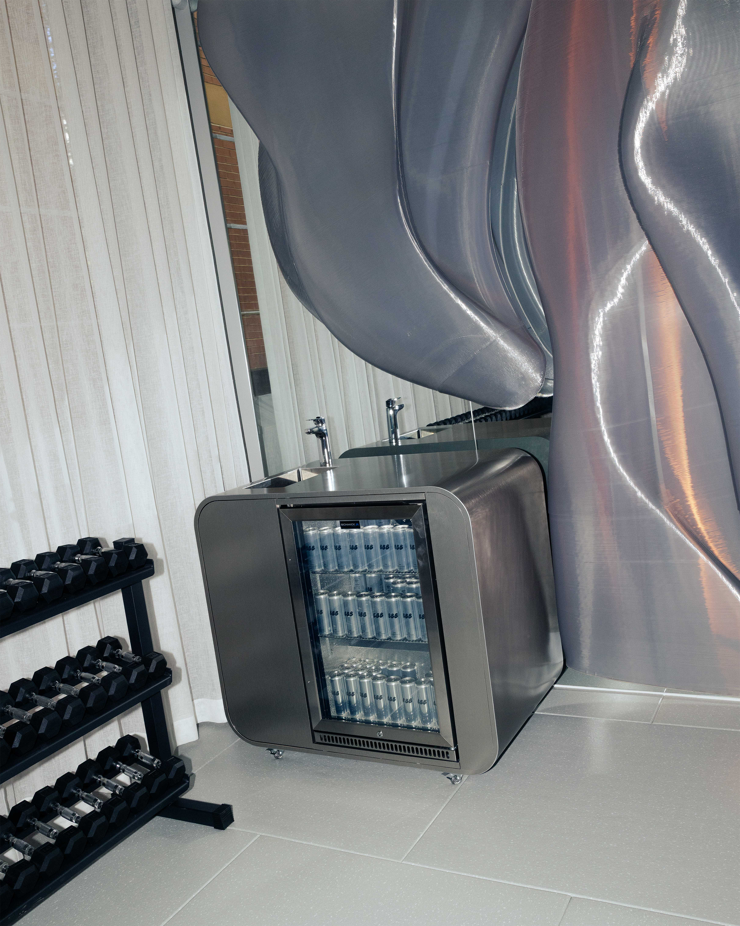 A gym or fitness area with a rack of black dumbbells on the left and a small refrigerator or cooler stocked with cans of water in the center. There is a large, metallic, curved object, possibly a piece of gym equipment, on the right side. The background includes light-colored wooden wall paneling and windows.