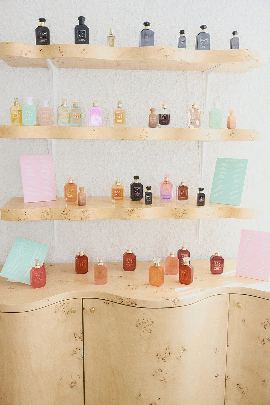 Display of KAYALI perfume bottles on wooden shelves with pink and green informational cards.