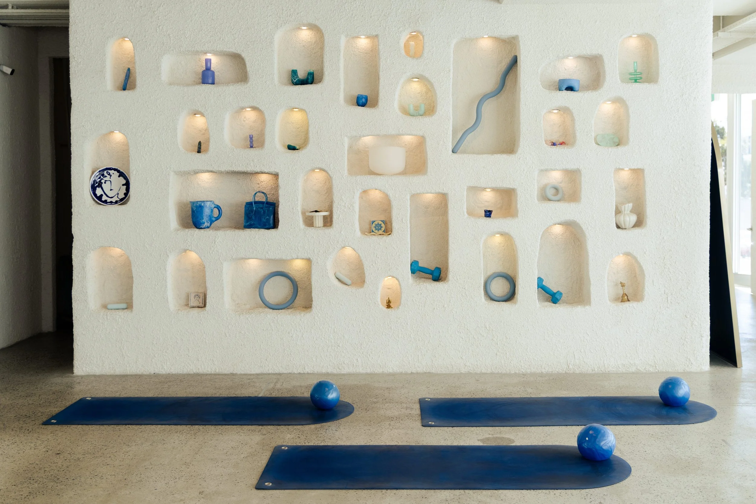 Yoga studio with a white textured wall and built-in lit niches displaying decorative items in blue, green, and white. Two yoga mats are on the beige carpeted floor, each with a blue exercise ball, and a few additional blue balls are scattered nearby.