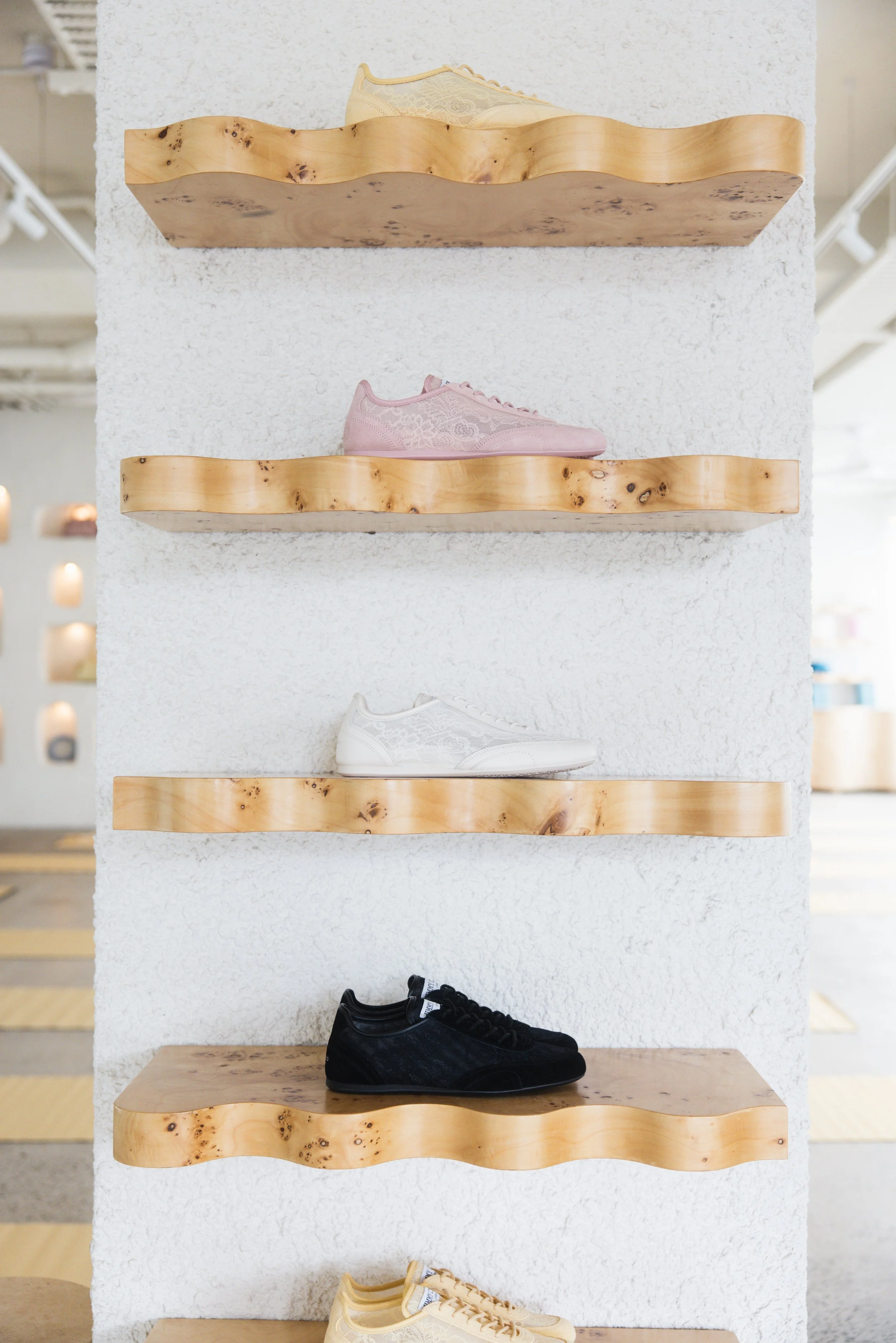 Display of sneakers on wooden shelves against a white textured wall, including beige, pink, black, and cream-colored shoes.
