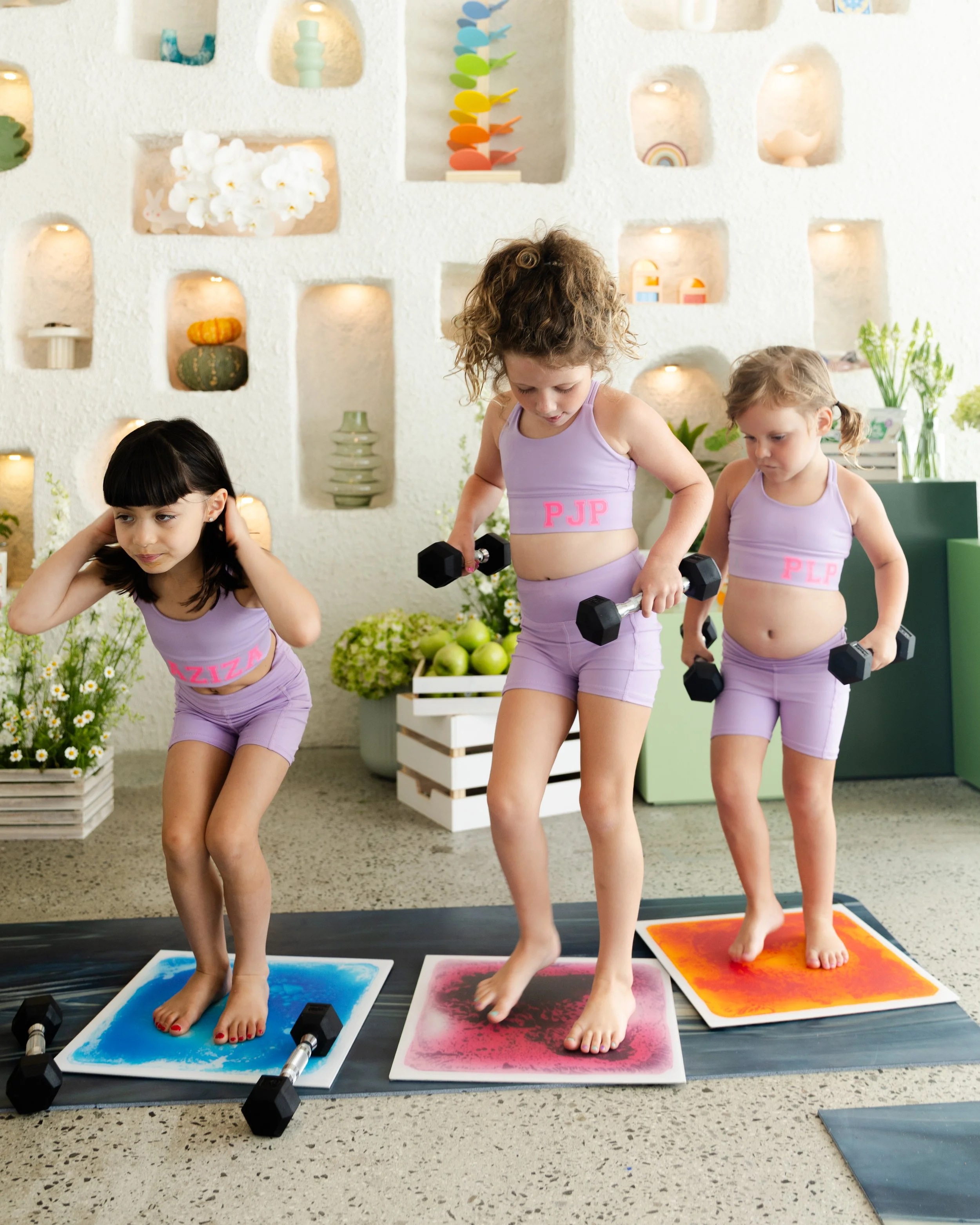 Three young girls in purple workout outfits doing a fitness class on colorful mats, lifting small dumbbells, in a bright room with decorative shelves and plants.